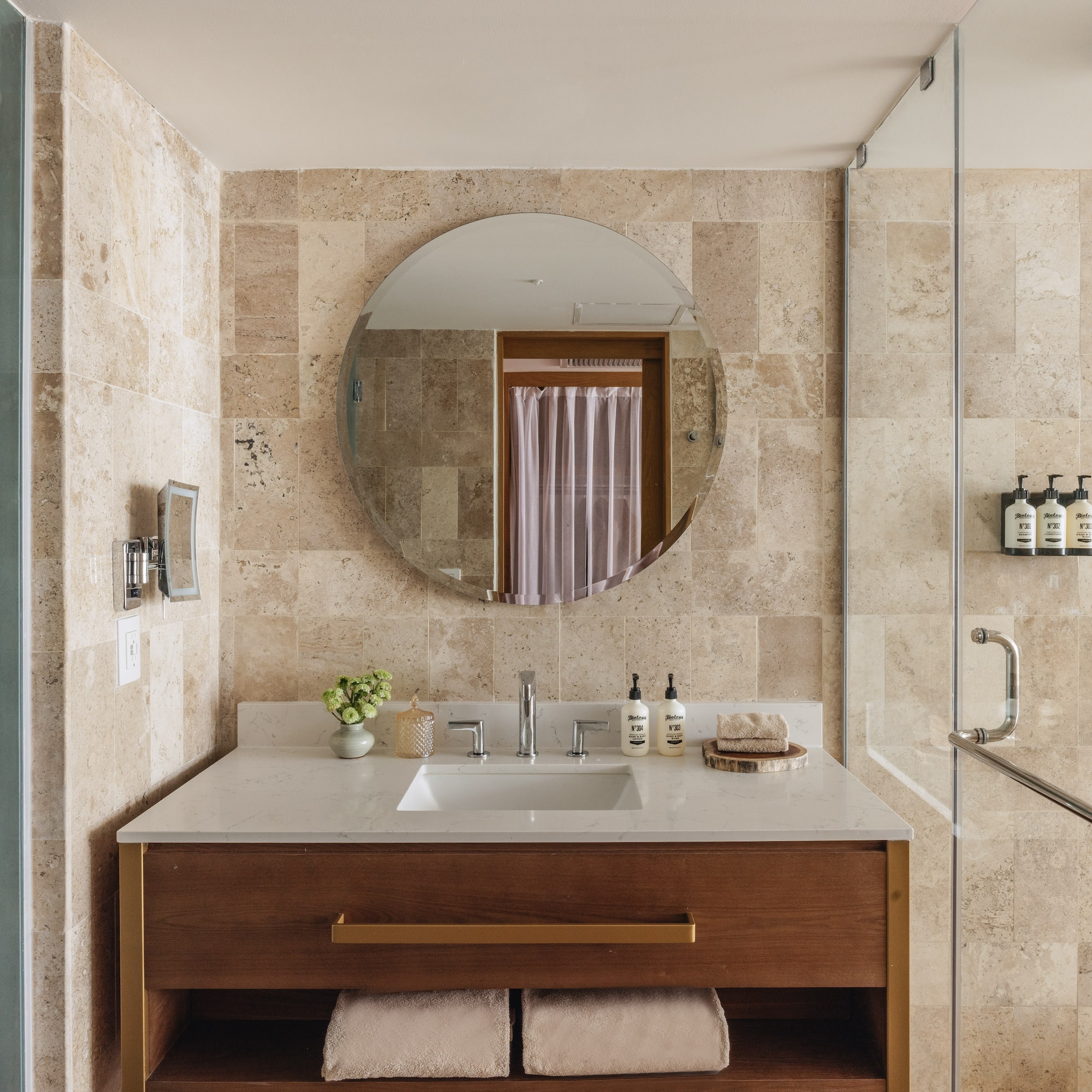 Modern hotel bathroom with wood vanity, travertine walls, and glass shower.