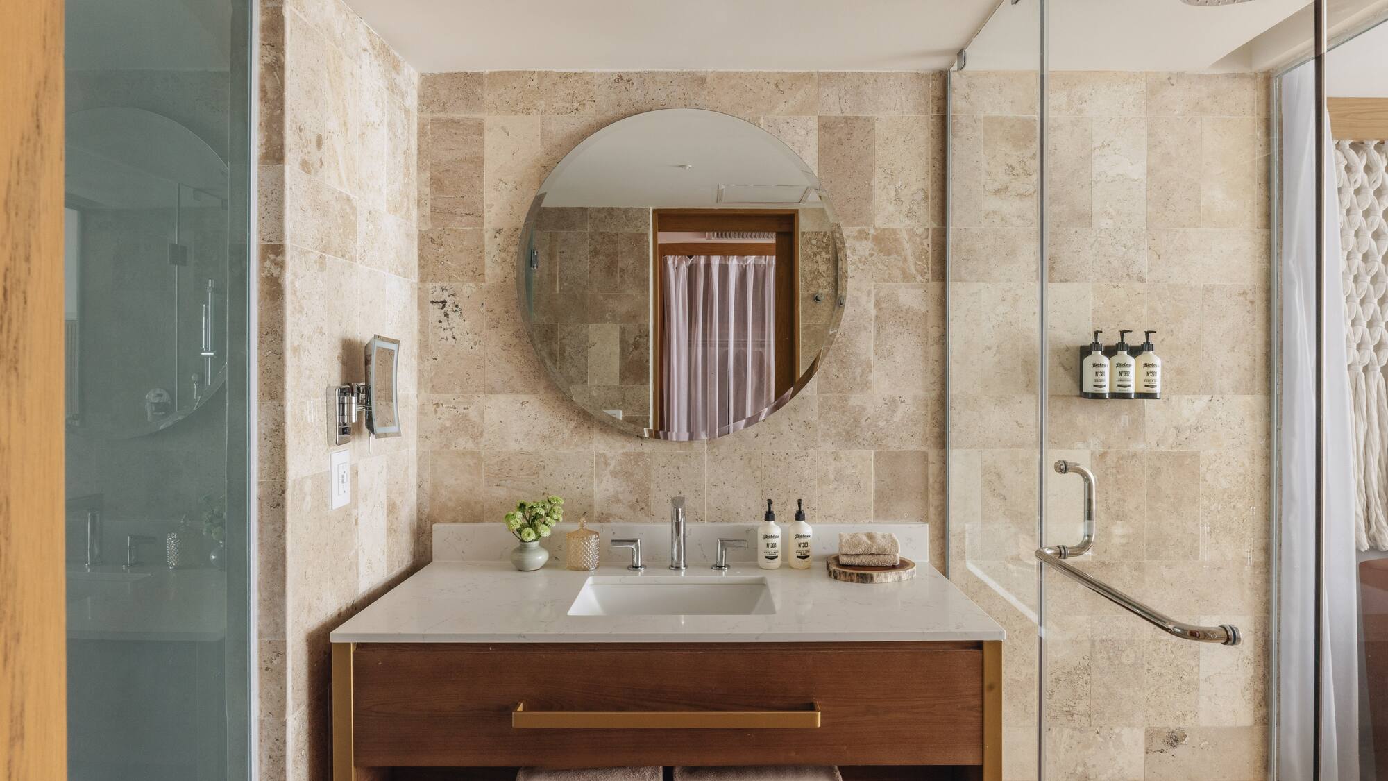 Modern hotel bathroom with wood vanity, travertine walls, and glass shower.