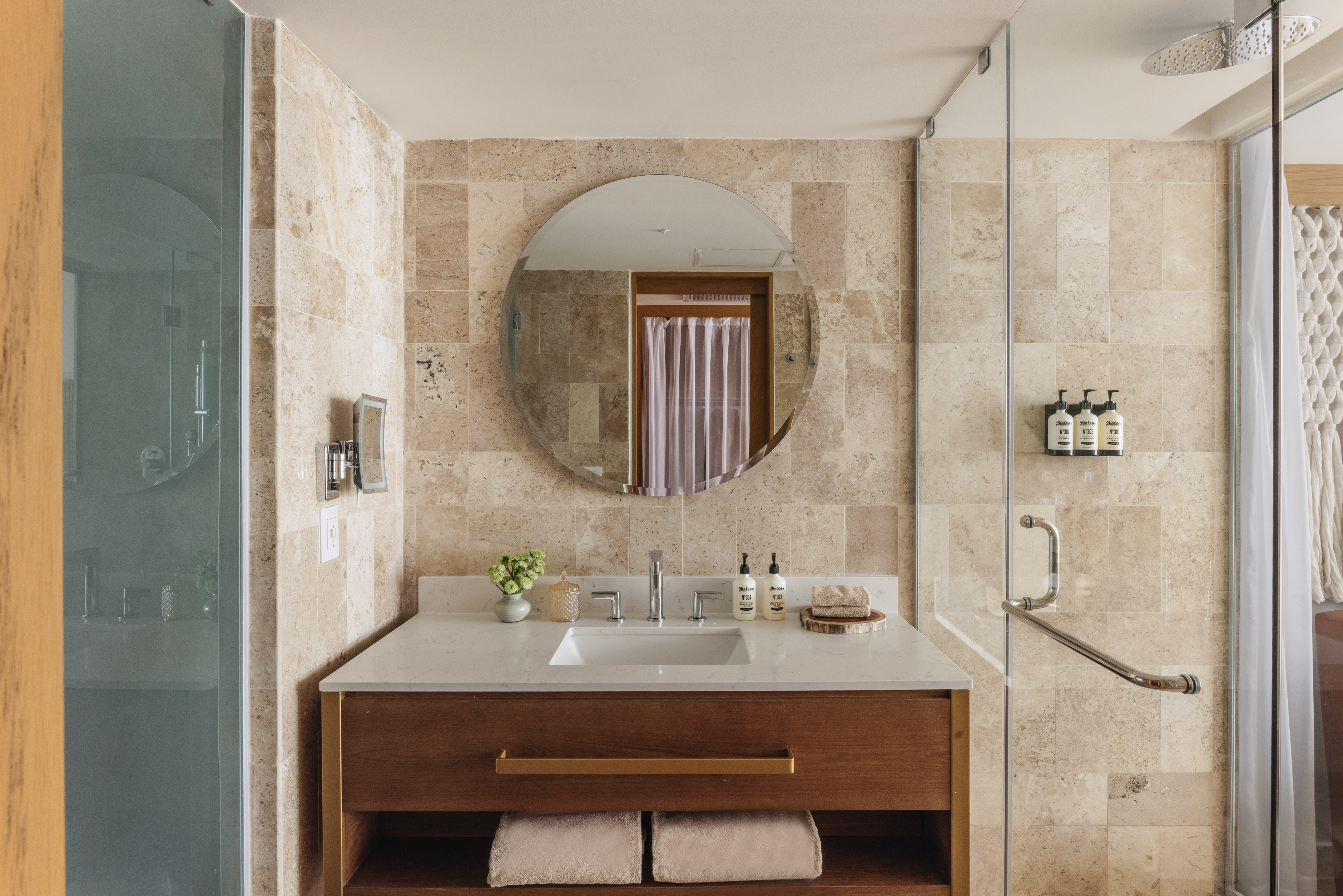 Modern hotel bathroom with wood vanity, travertine walls, and glass shower.