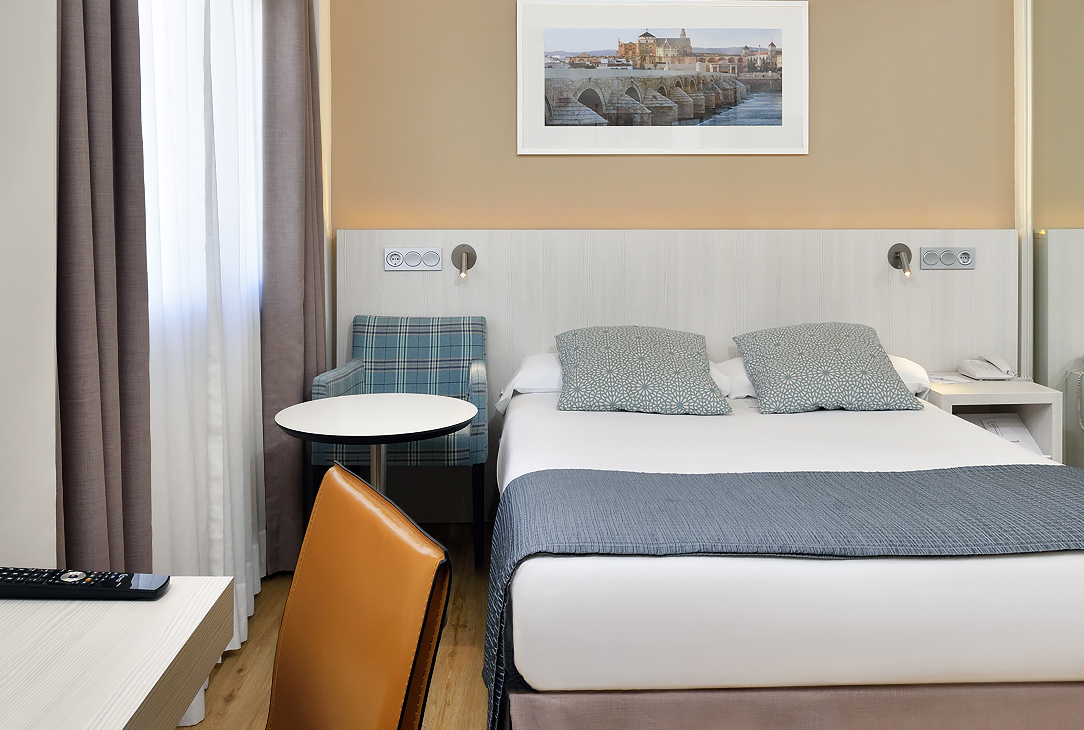 Modern hotel bedroom with bed, grey throw, plaid armchair, and bridge art.