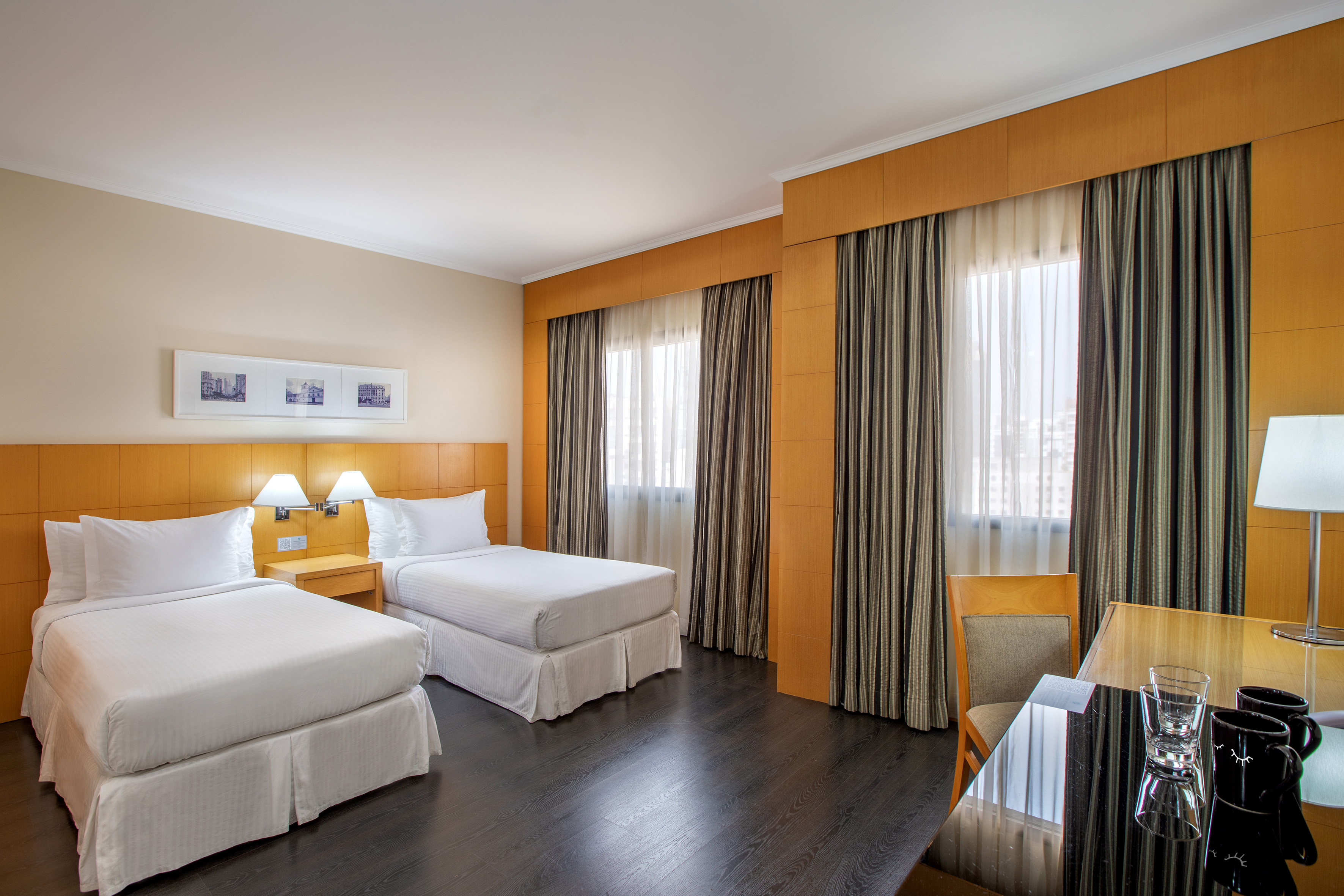 Modern hotel room with two twin beds, dark wood floor, and bright windows.