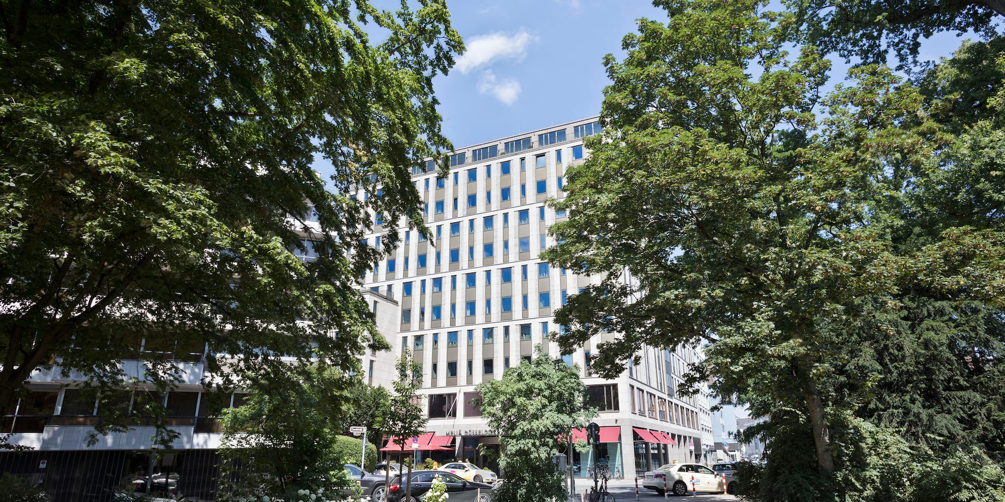 Tall modern building with many windows, framed by large green trees in a city.
