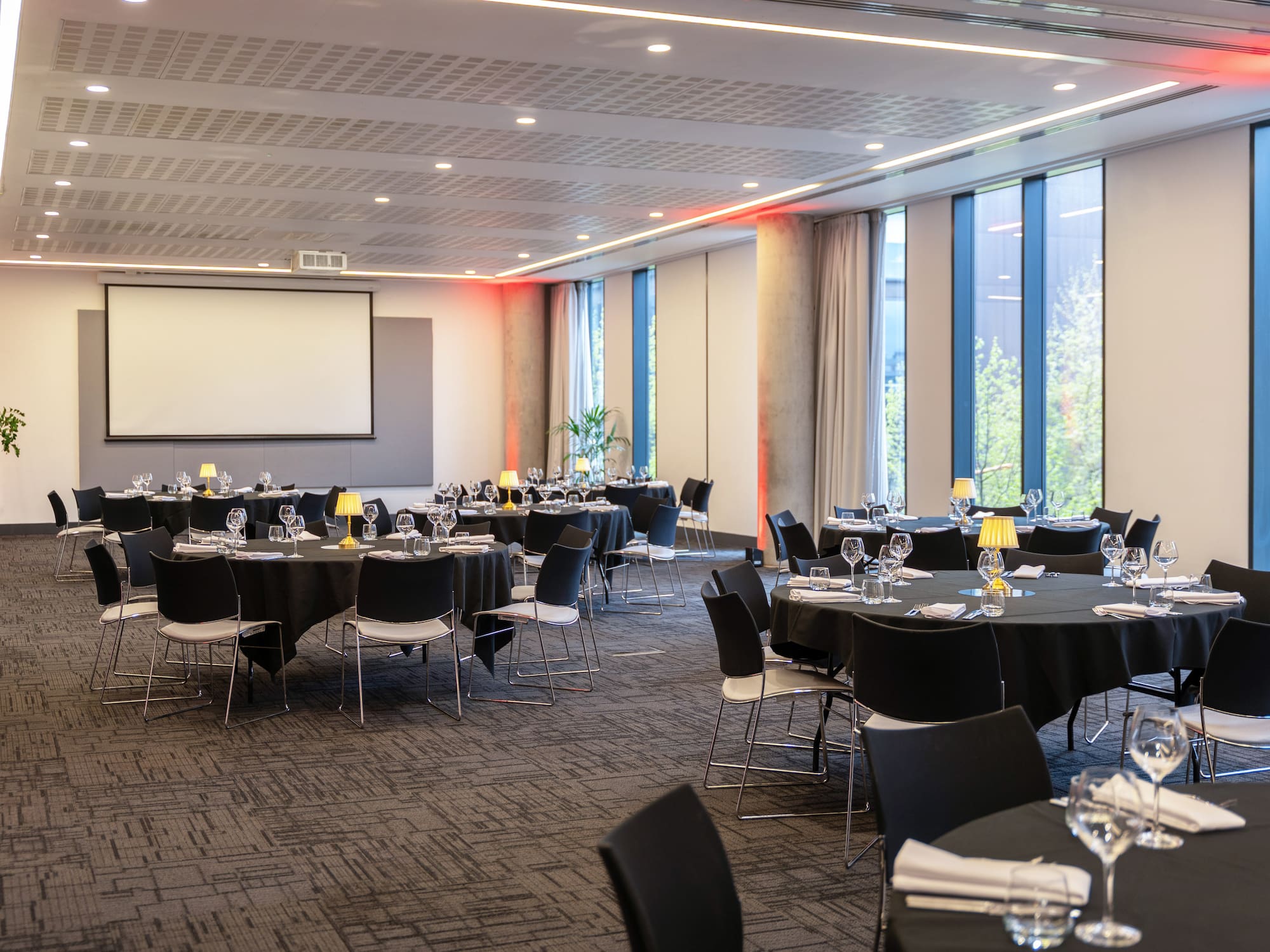 Modern event space with set round tables, screen, and red accent lighting.