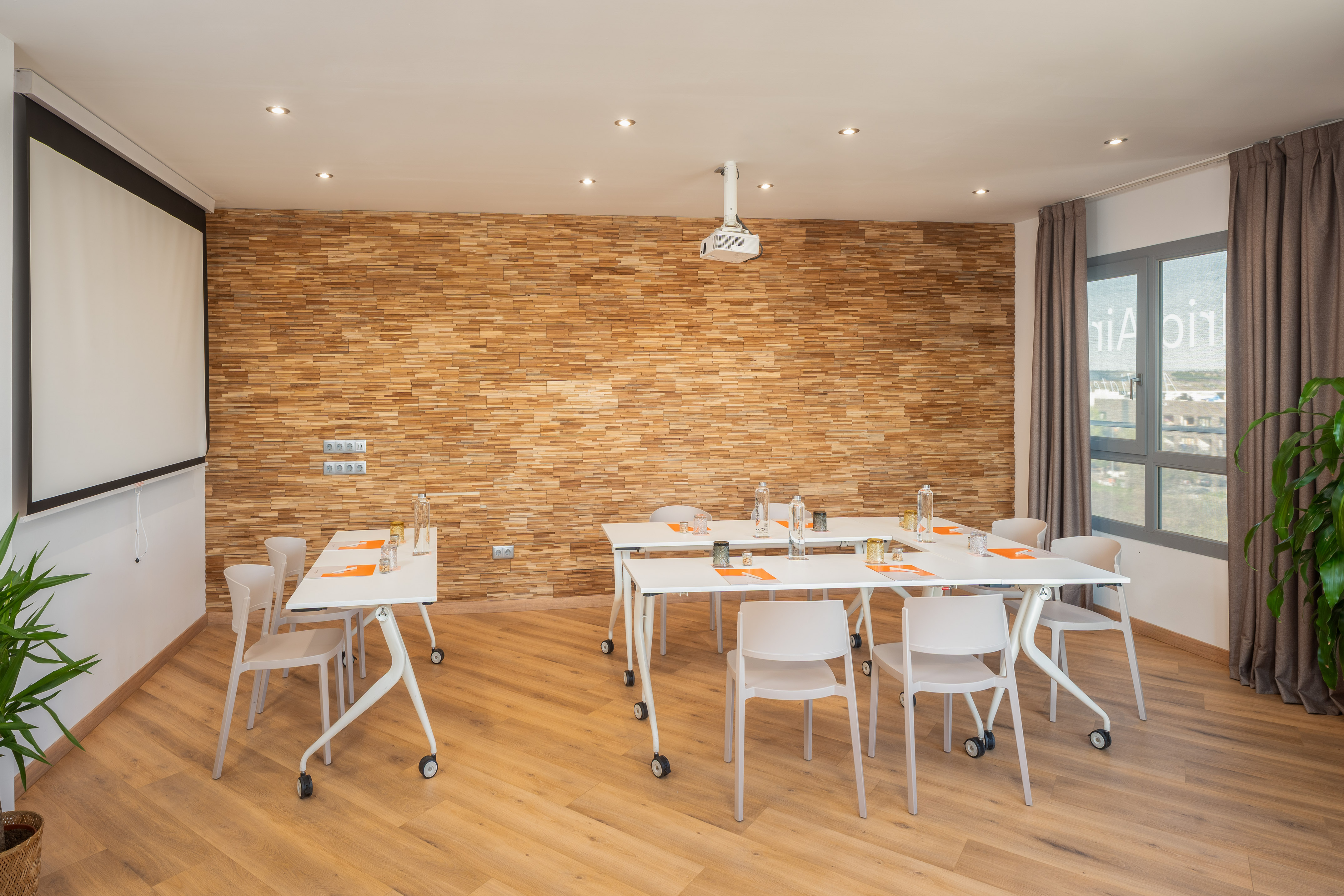 Modern meeting room with projector screen, white tables, and wood accent wall.