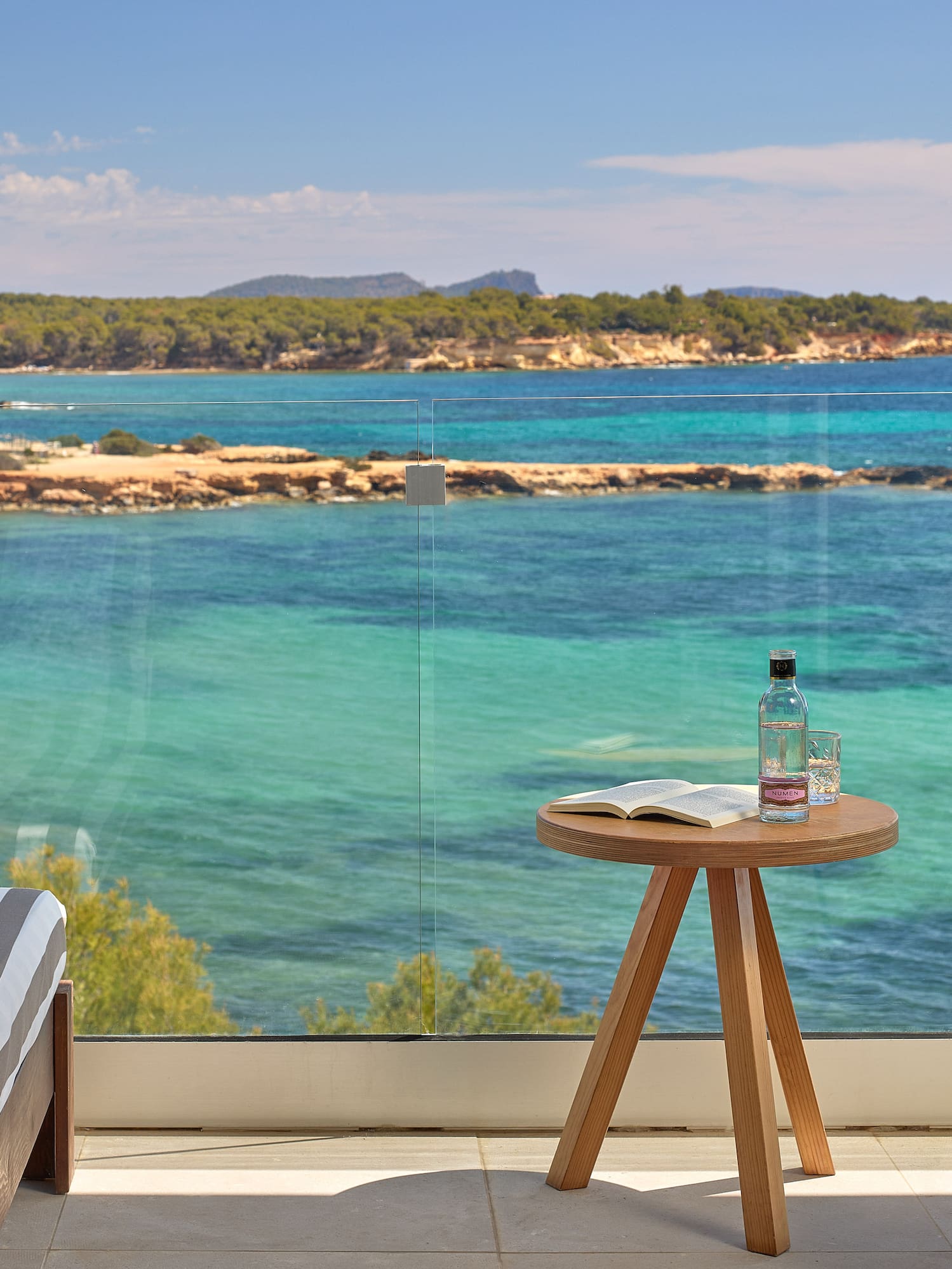 Hotel balcony with clear turquoise sea view, forested coast, and lounge chairs.