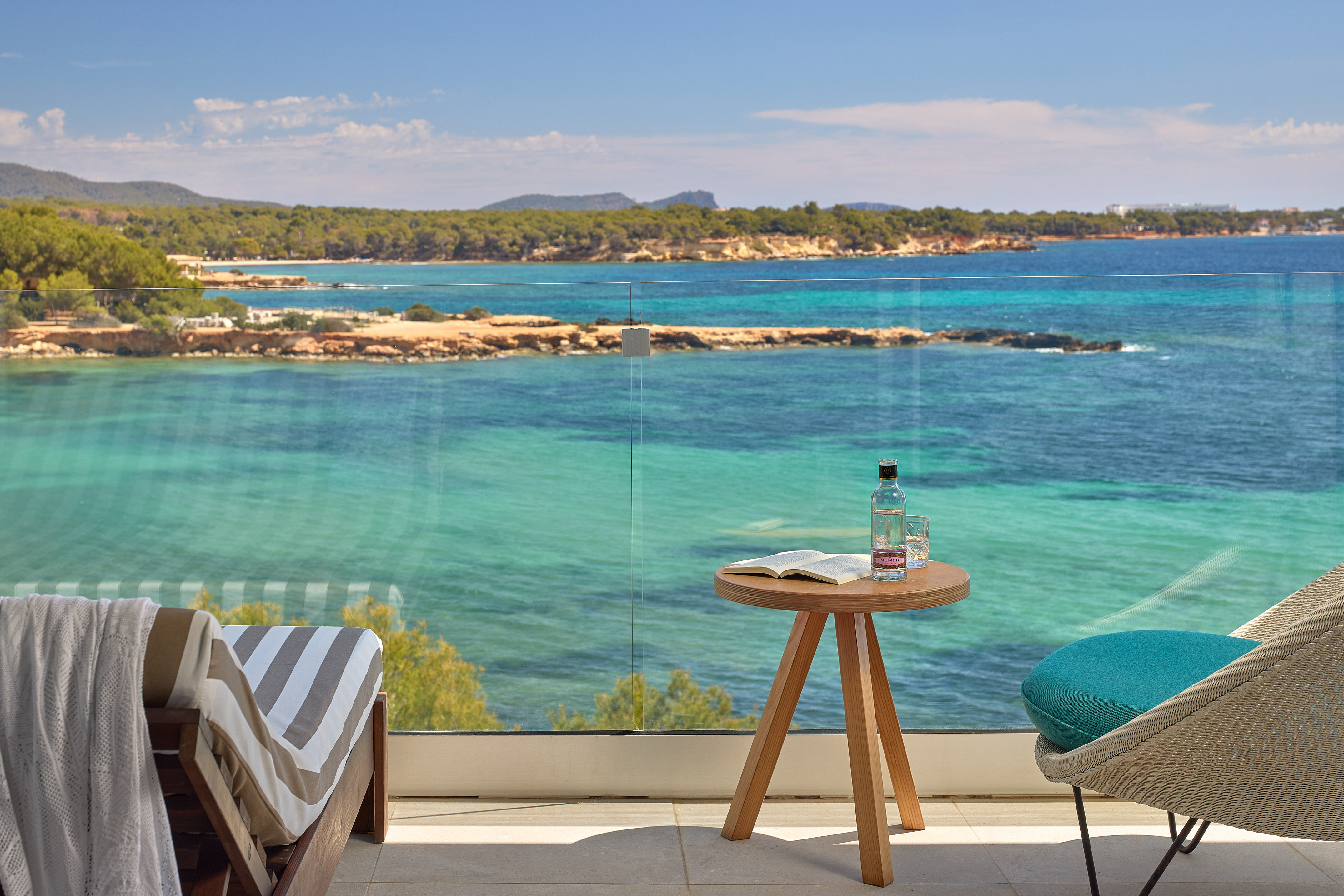 Hotel balcony with clear turquoise sea view, forested coast, and lounge chairs.