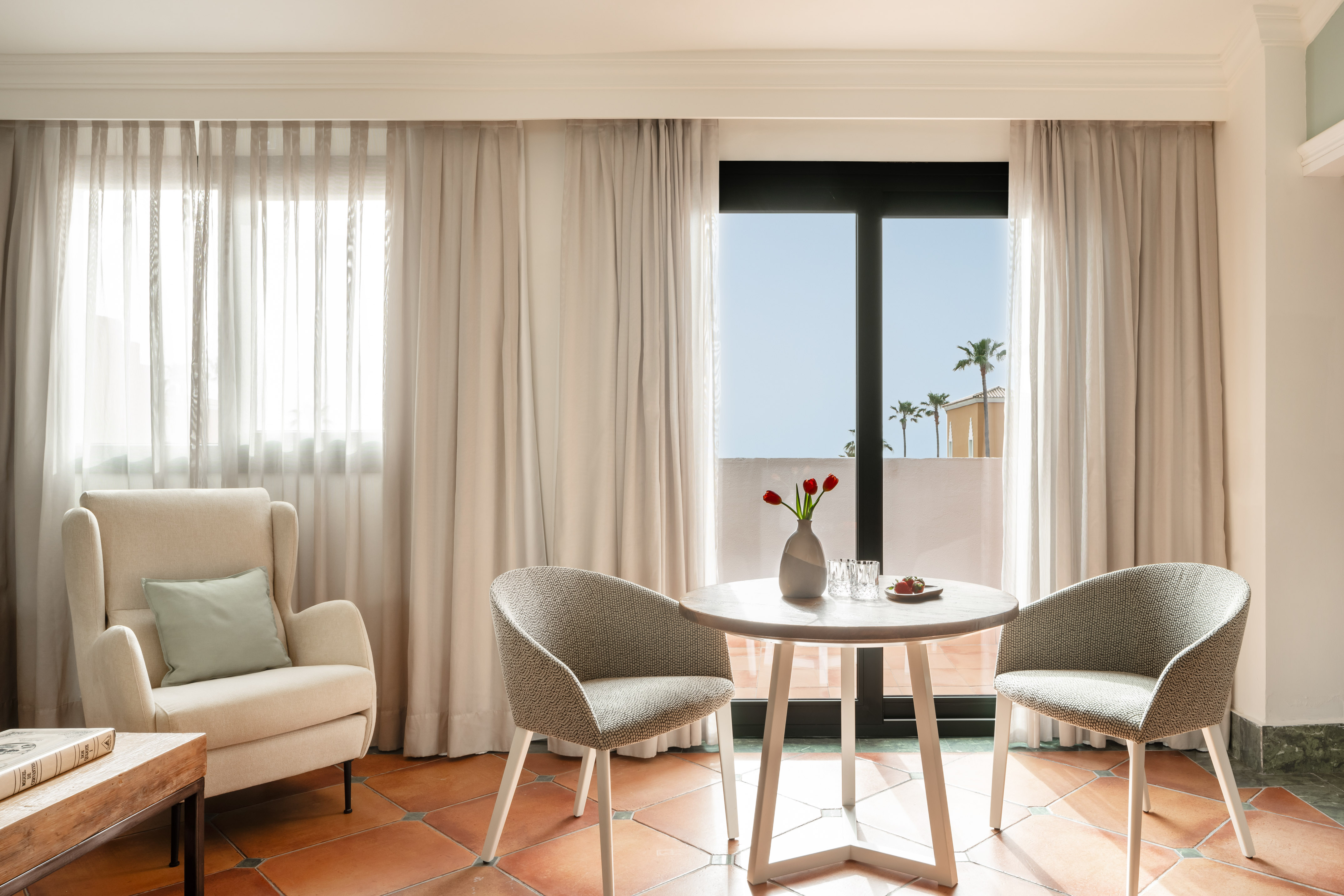 Modern hotel room with dining table, armchair, and balcony view.