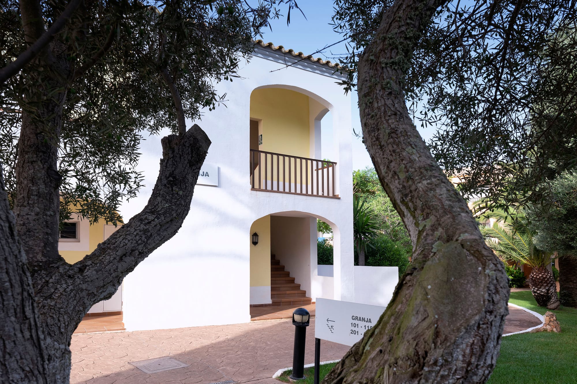 White Mediterranean resort building with balcony, yellow arches, olive trees.