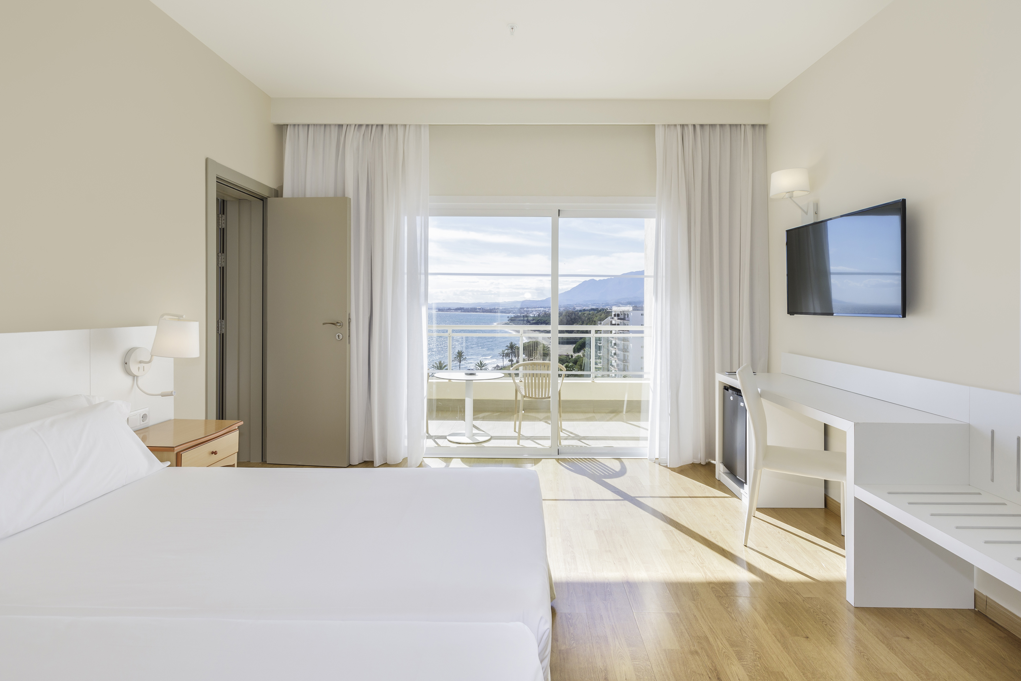 Modern hotel room with ocean view balcony, white bed, and TV.