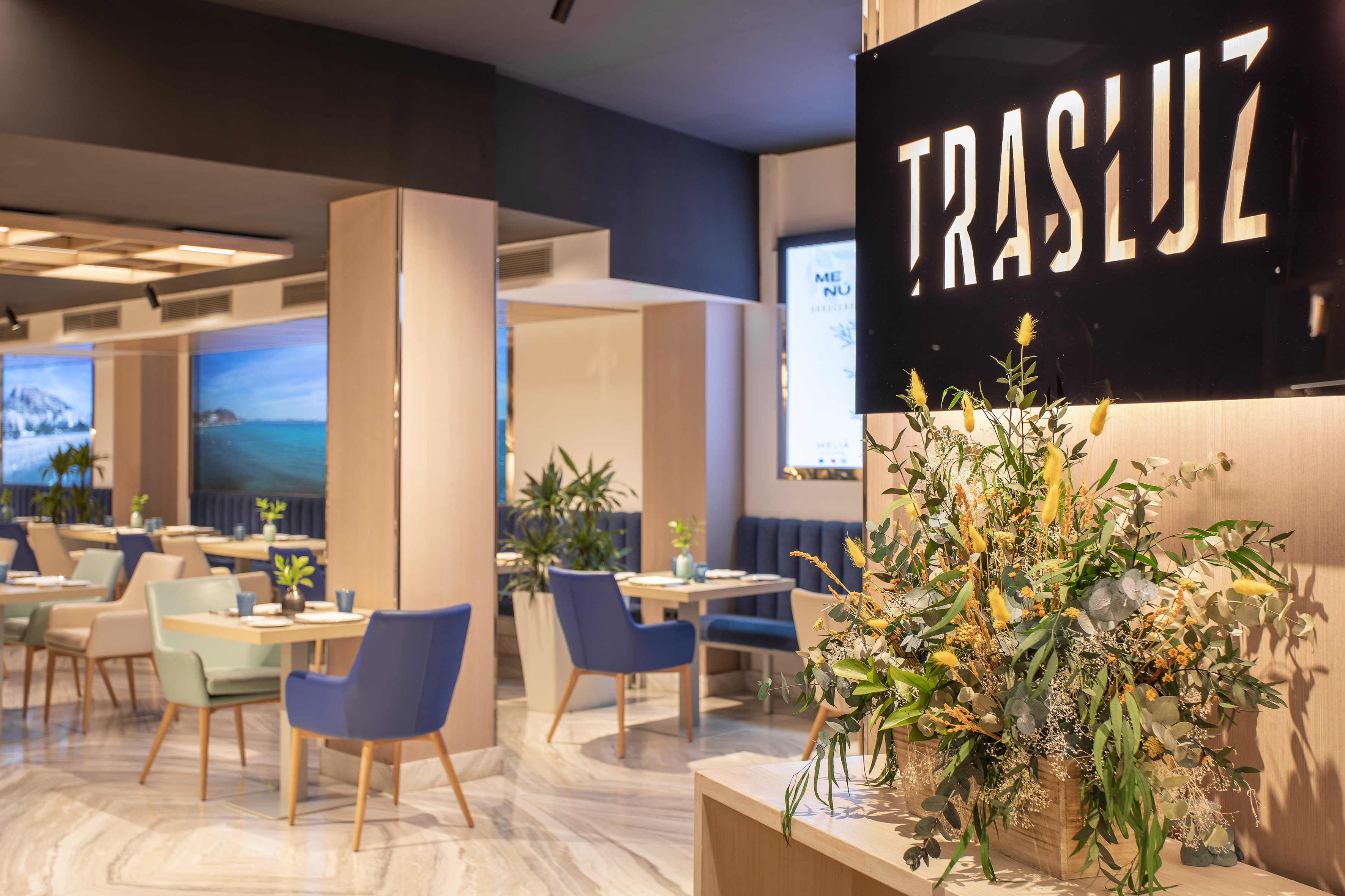 Modern Trasluz restaurant interior with tables, blue chairs, and floral display.