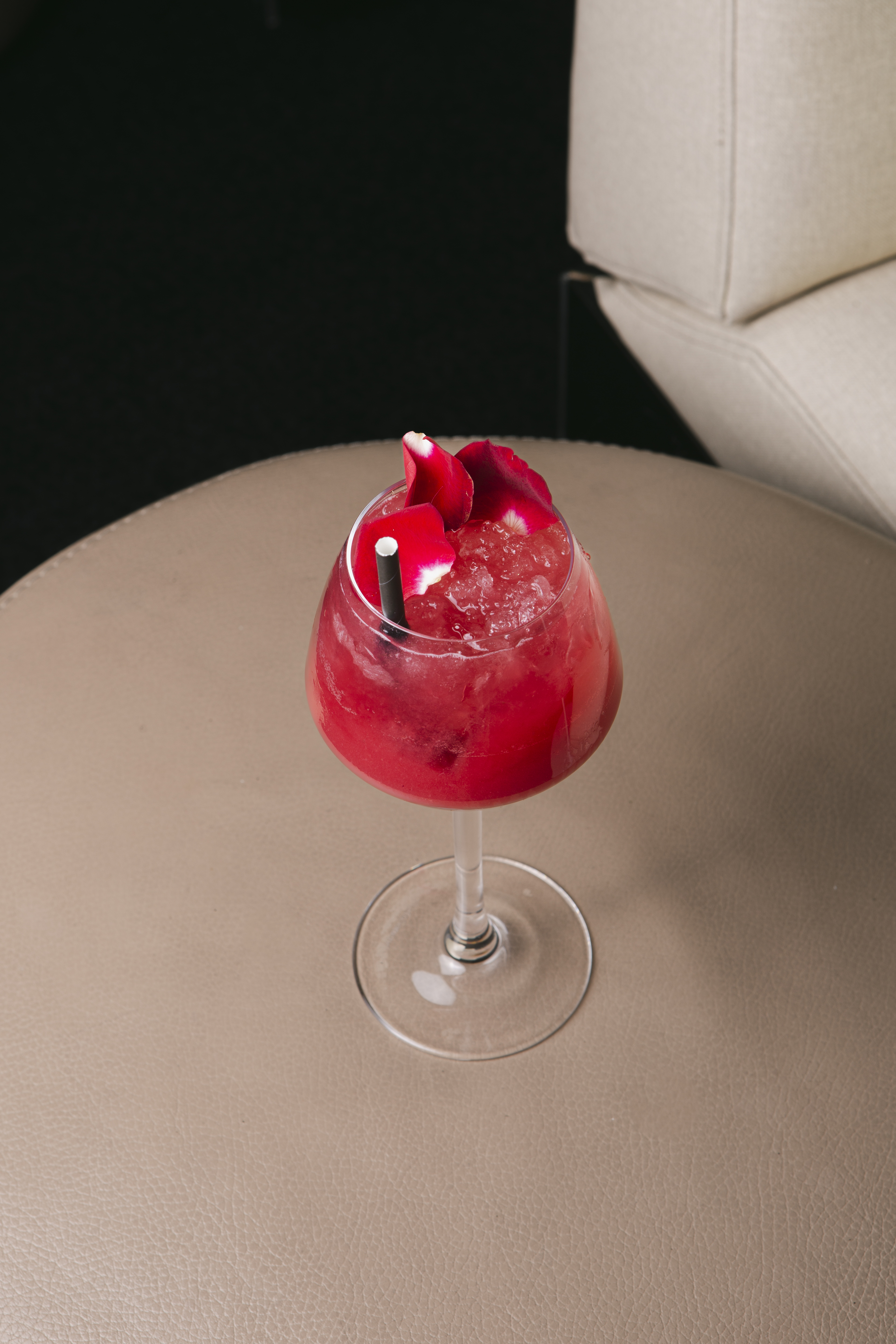Red cocktail with rose petals, crushed ice, and straw on a lounge table.