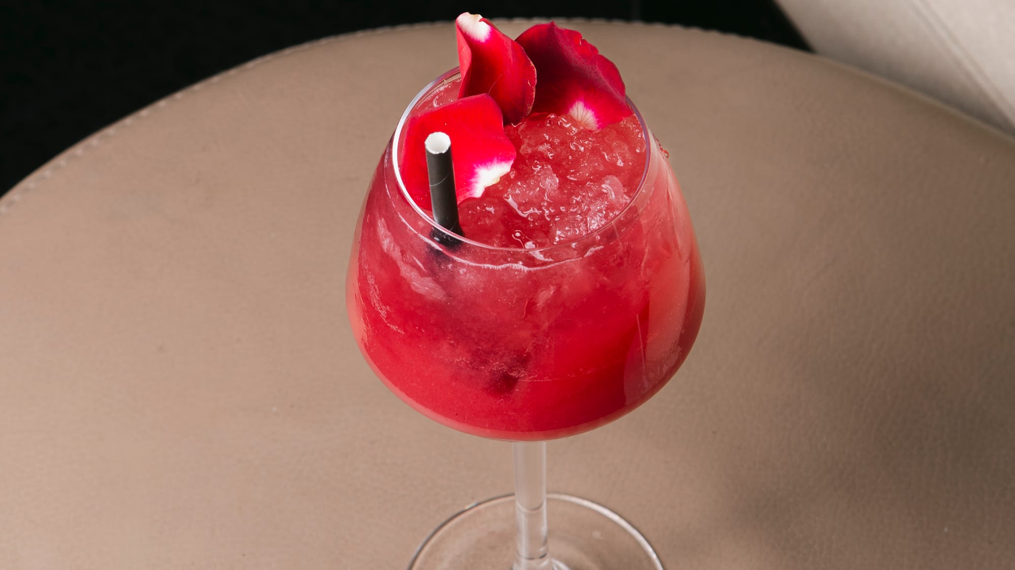 Red cocktail with rose petals, crushed ice, and straw on a lounge table.