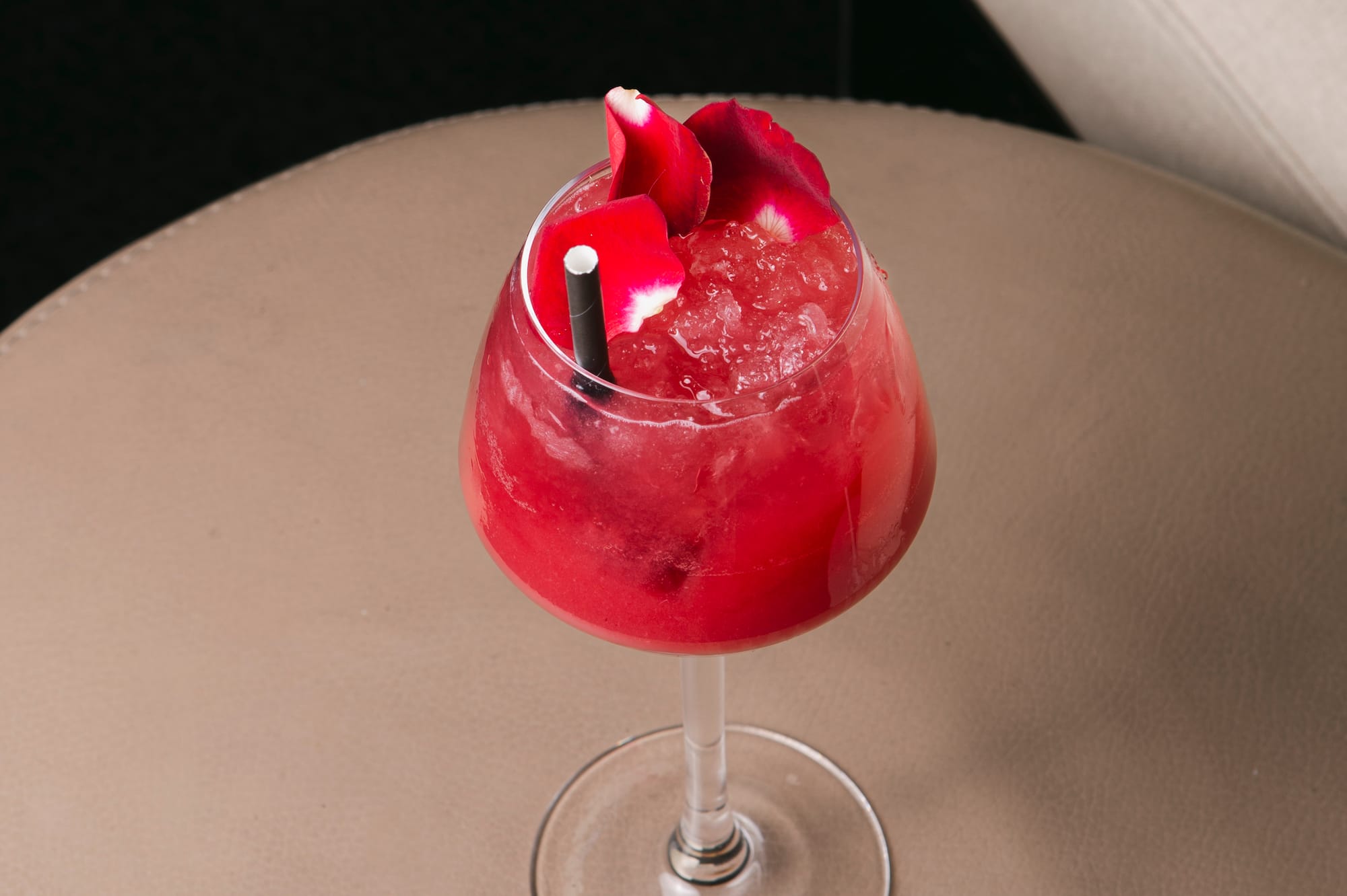 Red cocktail with rose petals, crushed ice, and straw on a lounge table.