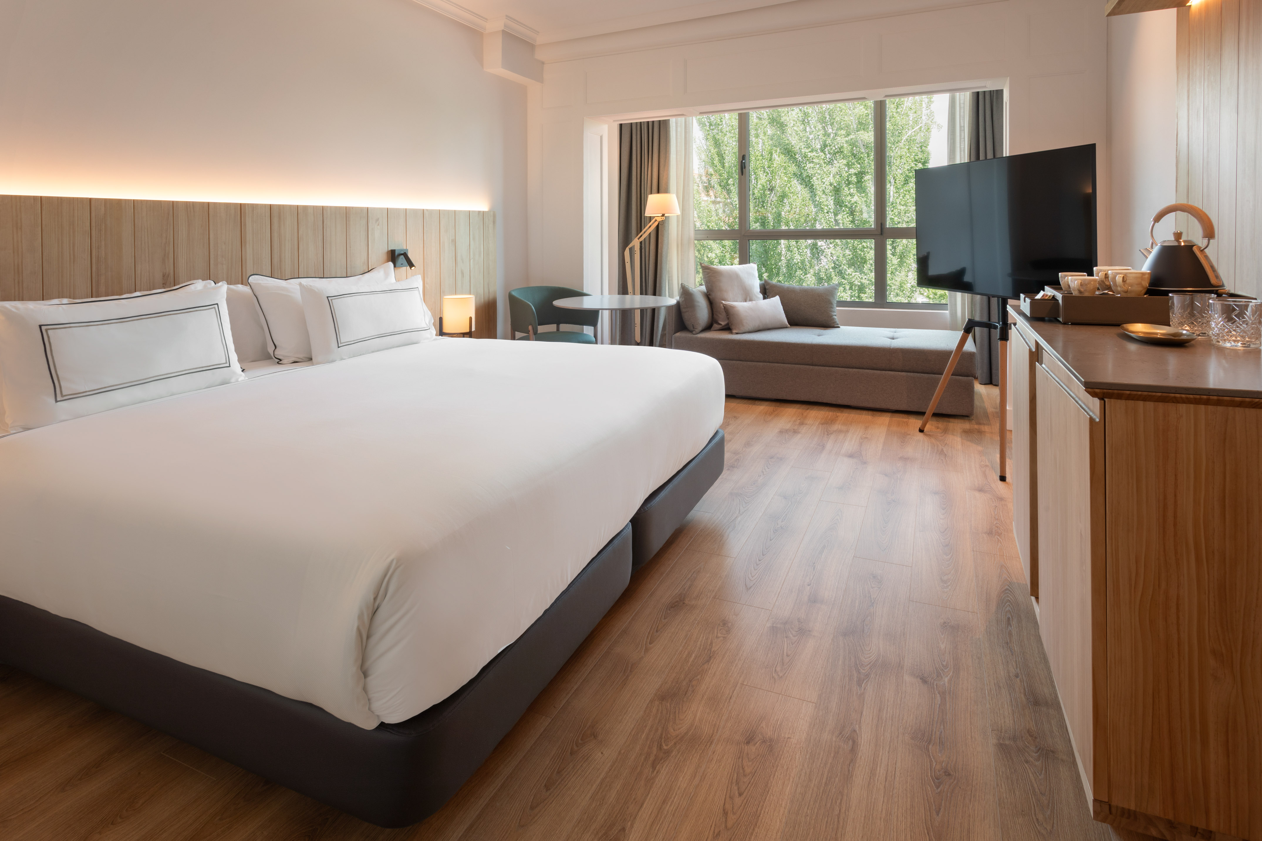 Modern hotel room with king bed, wooden headboard, window lounge, TV, and minibar.