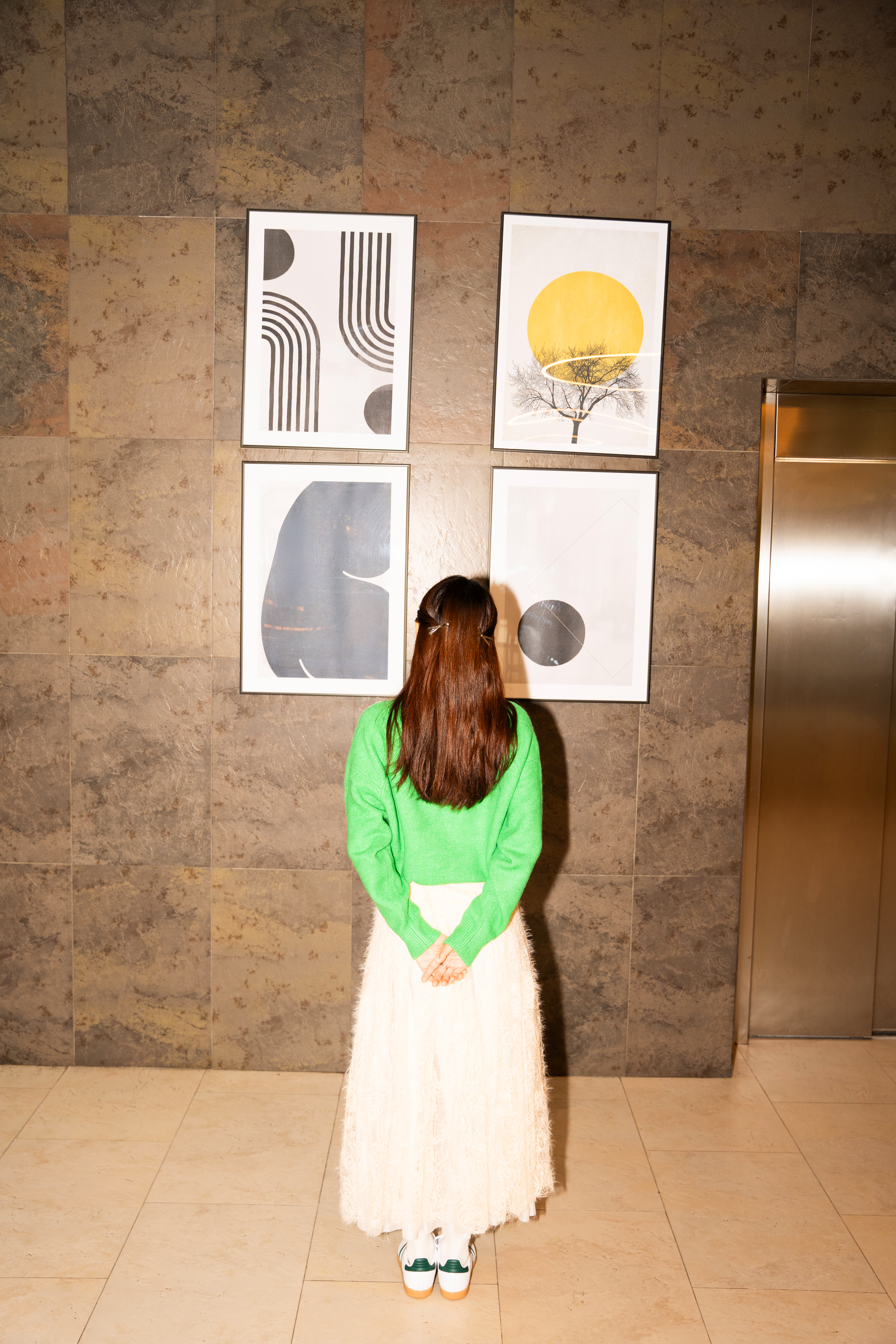 Woman in green sweater, cream skirt, viewing modern abstract art wall.