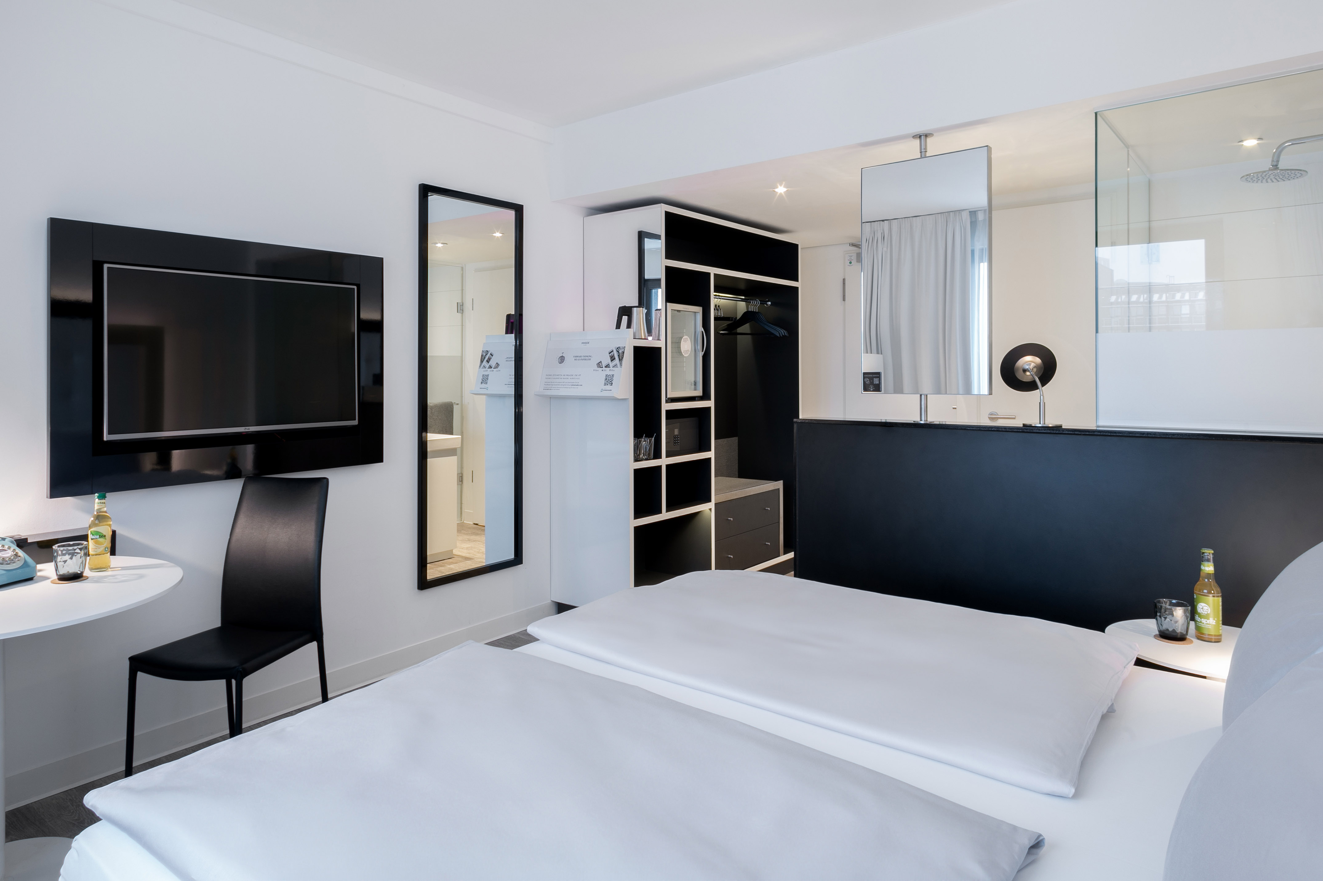 Modern white and black hotel room with bed, TV, wardrobe, and glass shower.