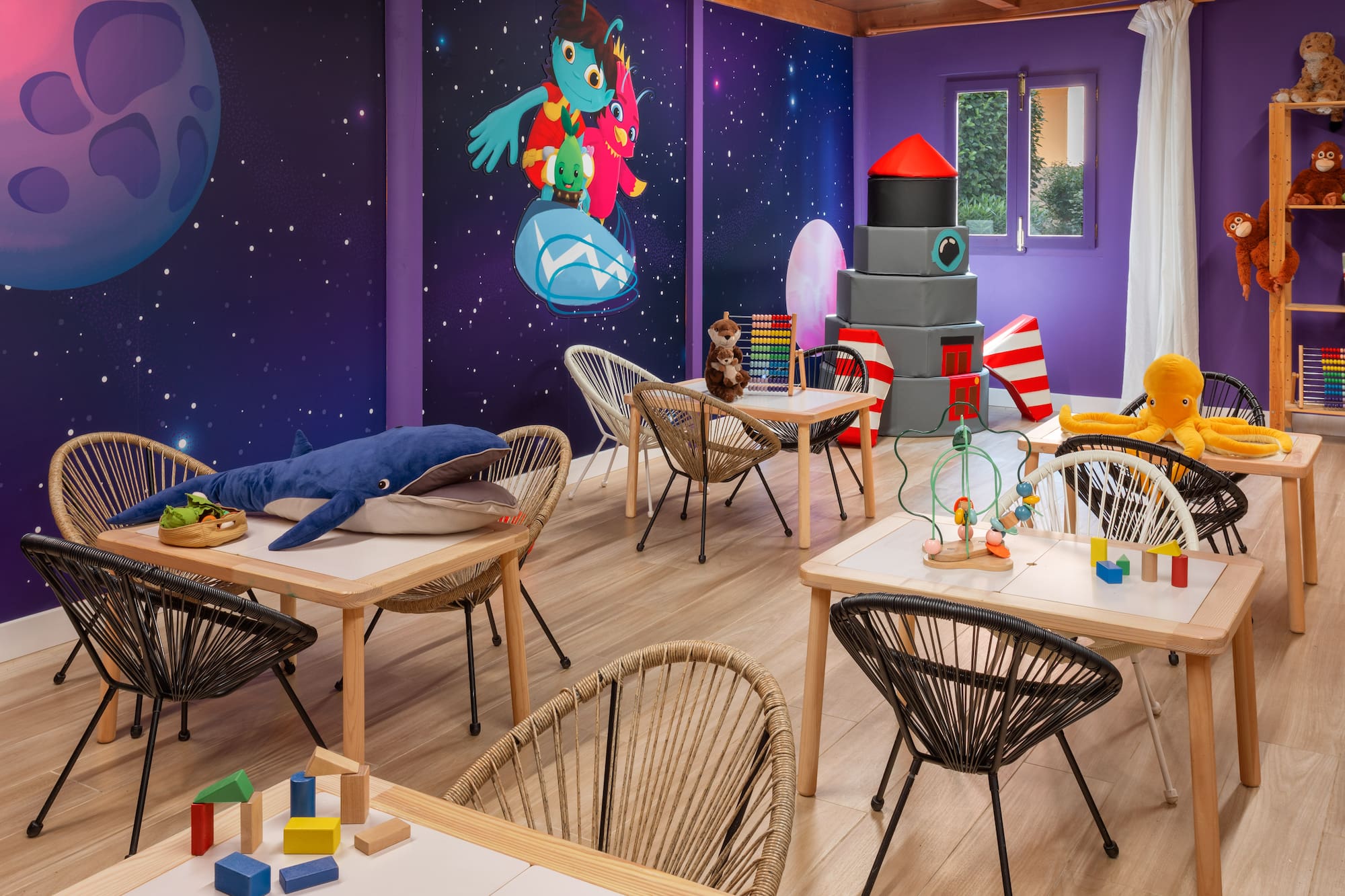 Kids' space playroom with mural, tables, chairs, building blocks, and plush toys.