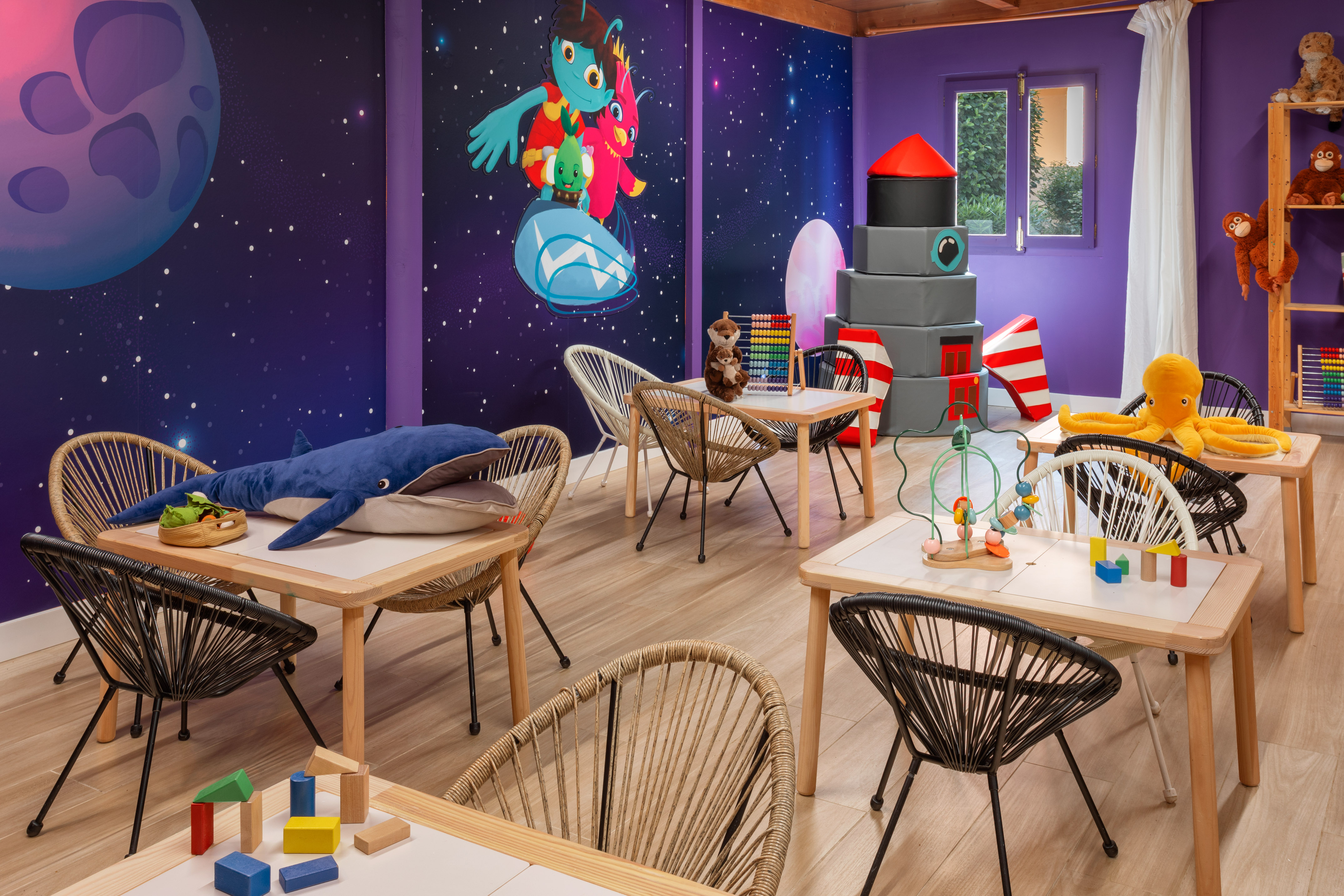 Kids' space playroom with mural, tables, chairs, building blocks, and plush toys.