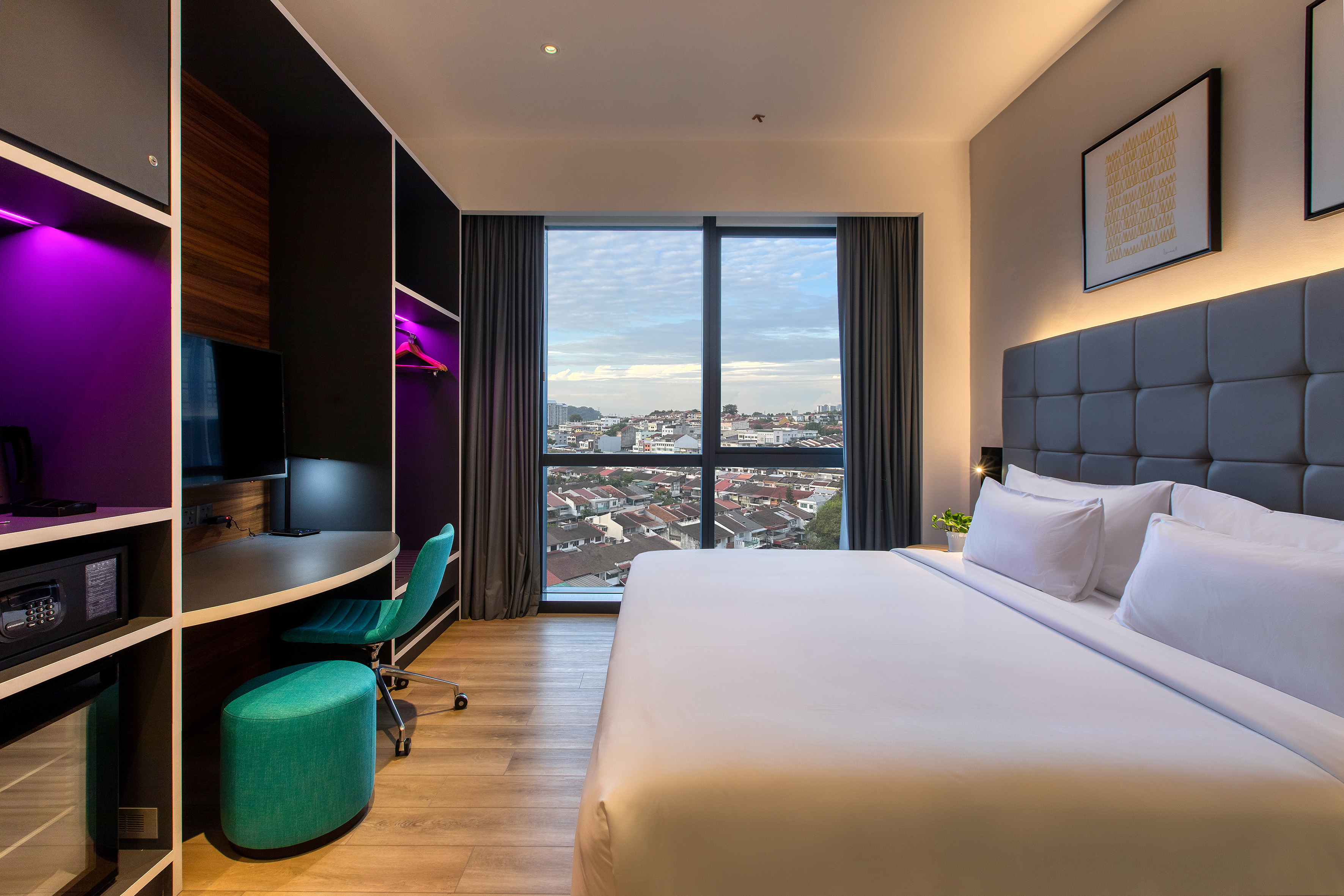 Modern hotel room with king bed, city view window, workspace, purple lighting.