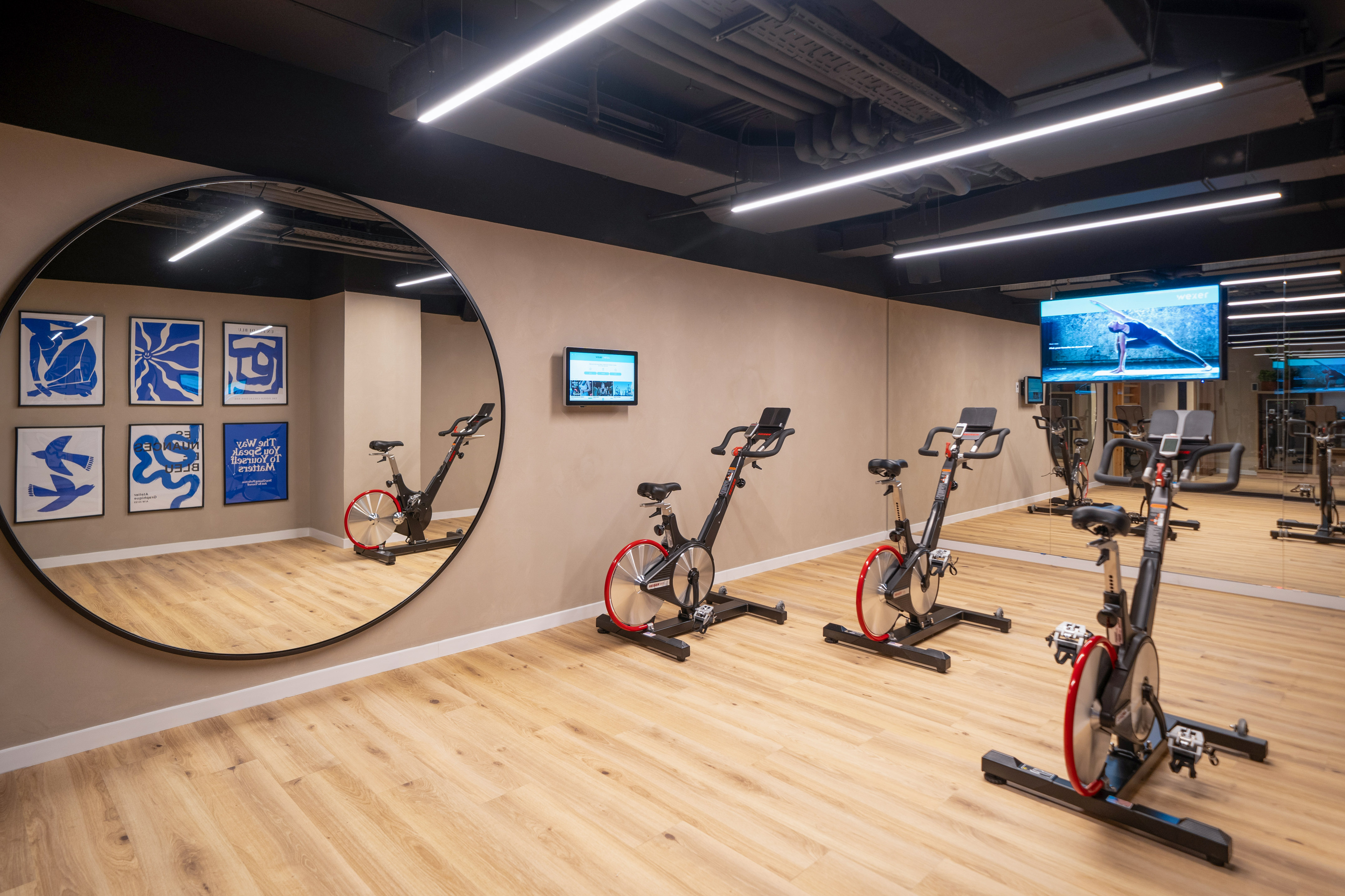 Modern gym with spin bikes, light wood floor, large mirrors, and wall displays.