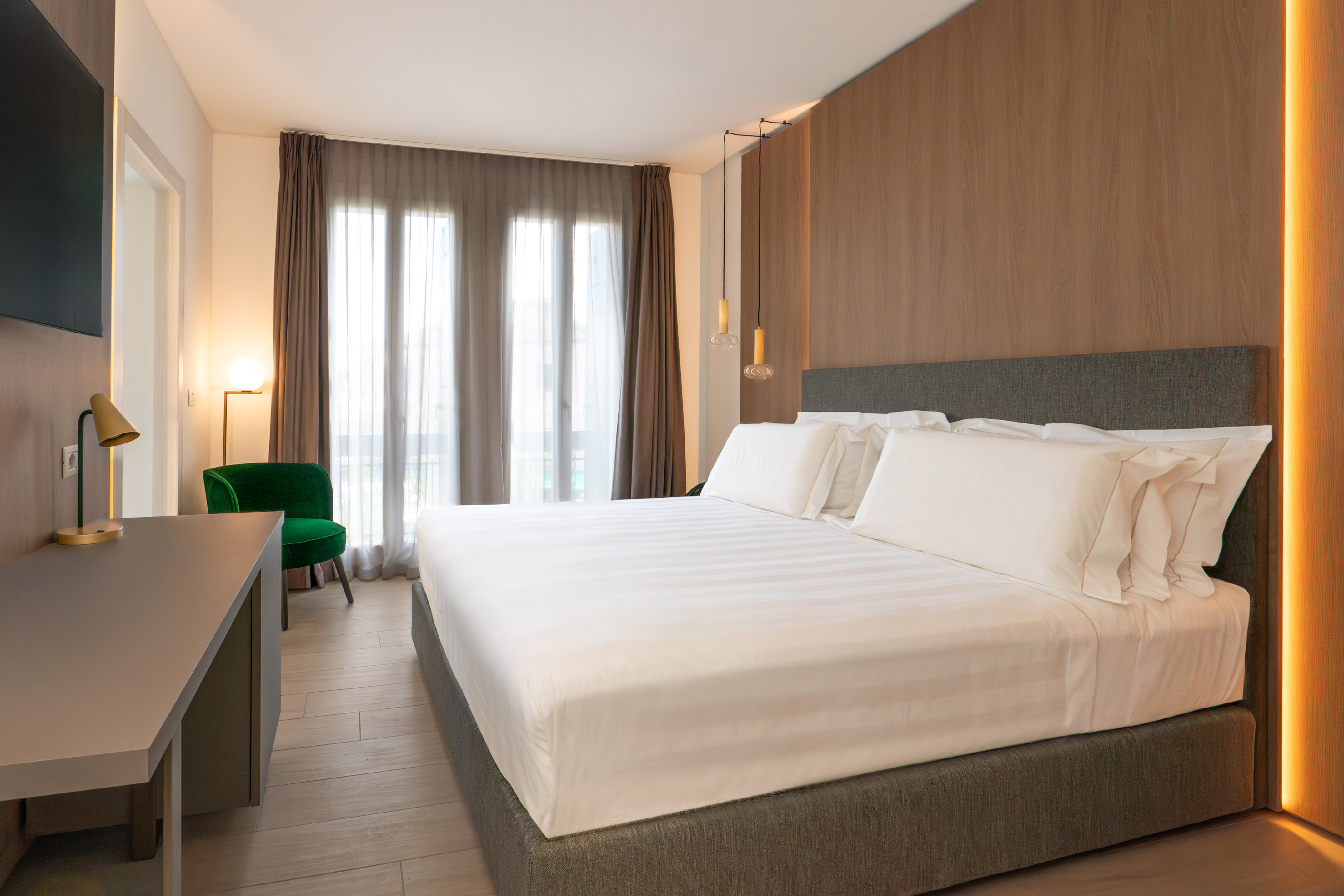 Modern hotel bedroom with king bed, wood wall, green armchair, and large windows.