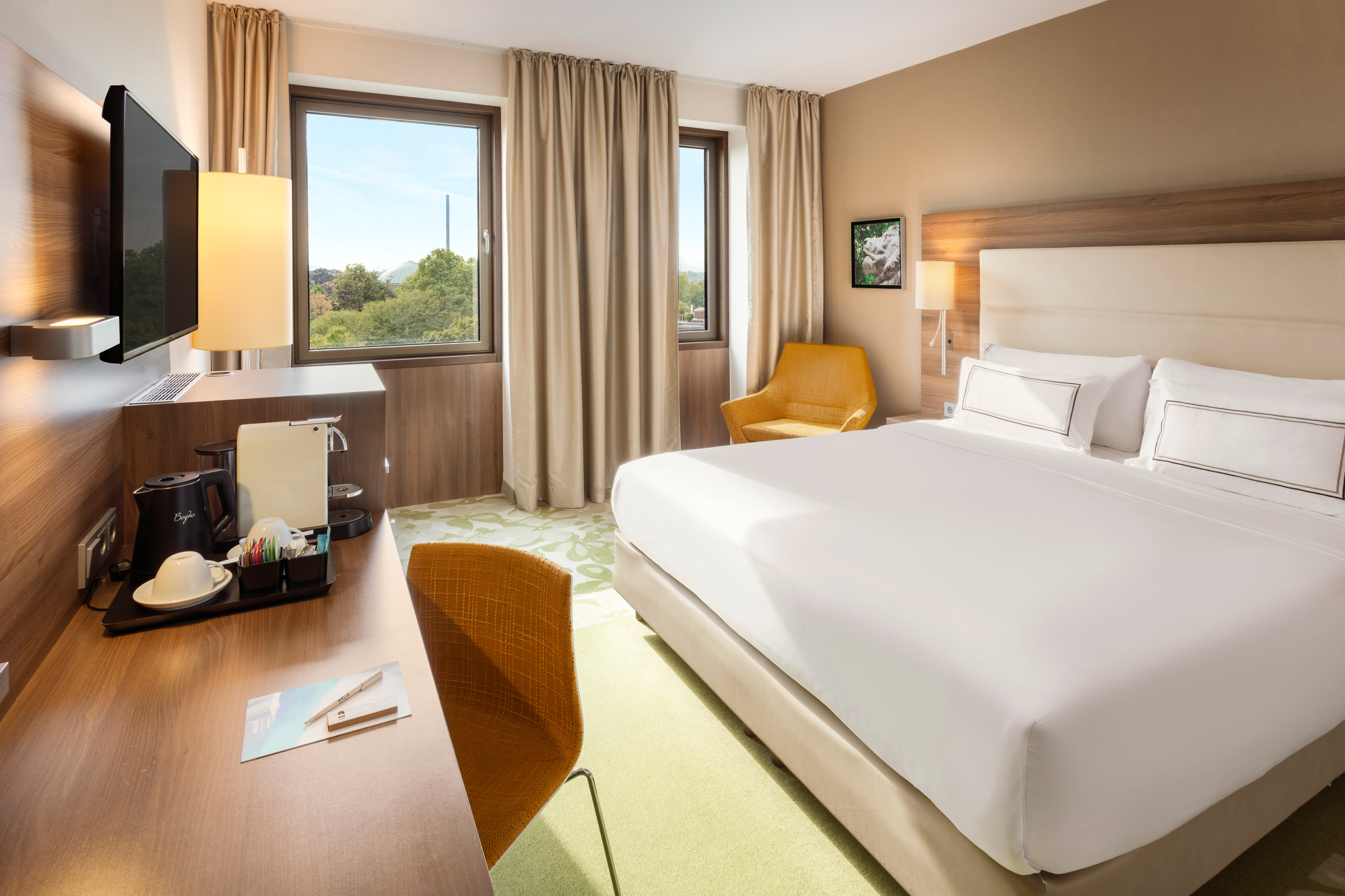 Bright hotel room with king bed, desk, TV, coffee maker, and window.