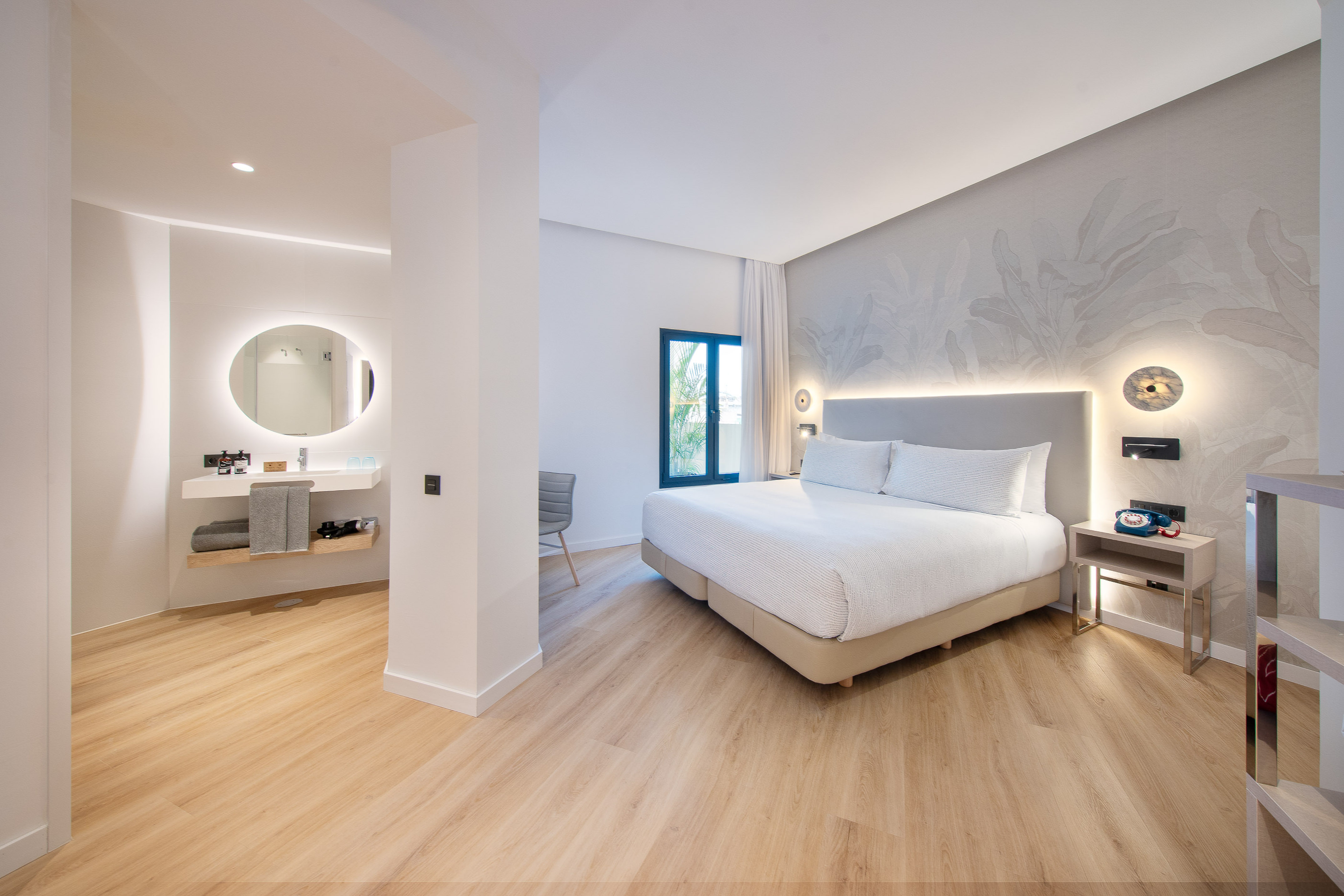 Modern hotel bedroom with queen bed, leaf wallpaper, wood floor, and open bathroom area.