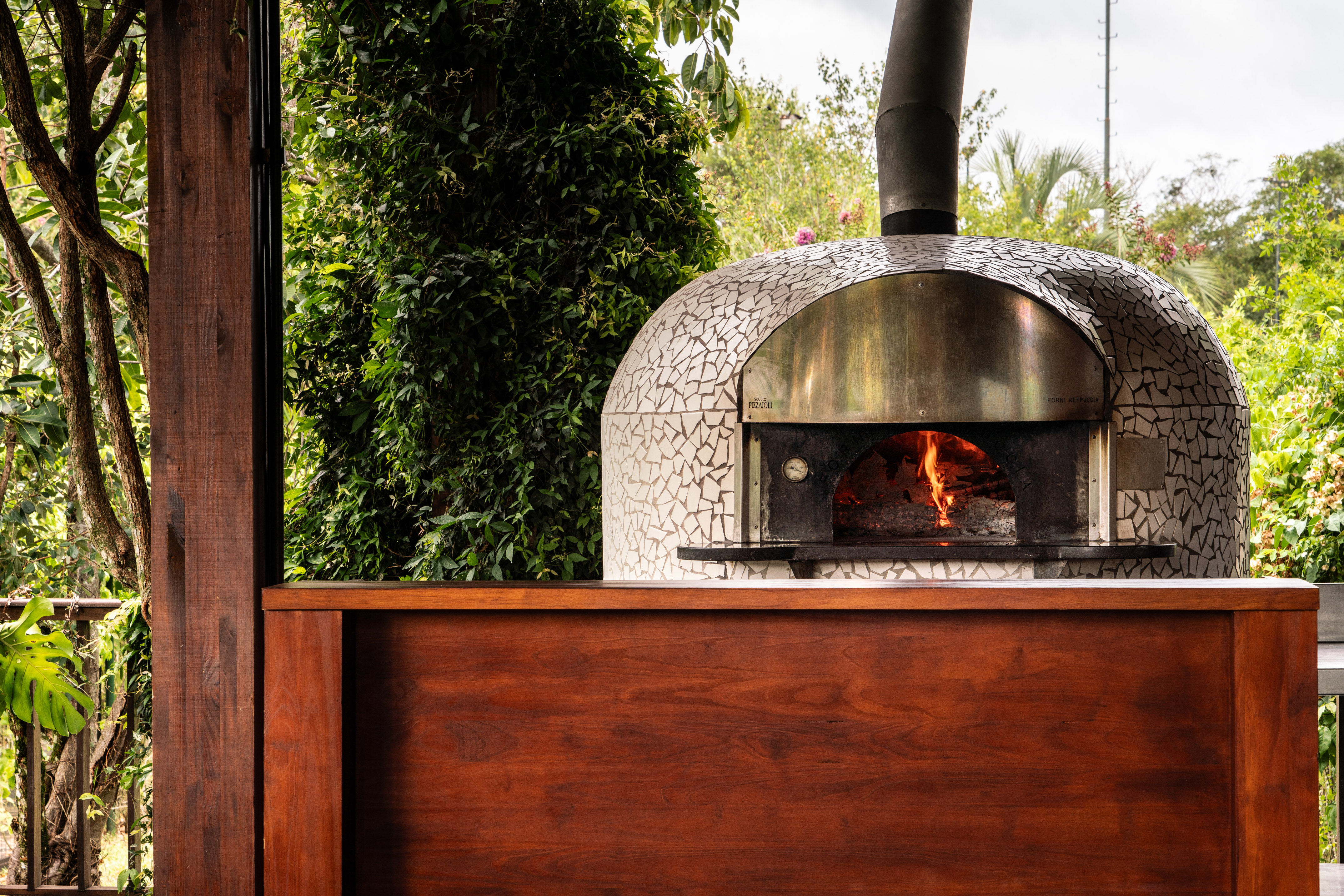 Mosaic tiled outdoor pizza oven with fire, wooden counter, and garden backdrop.