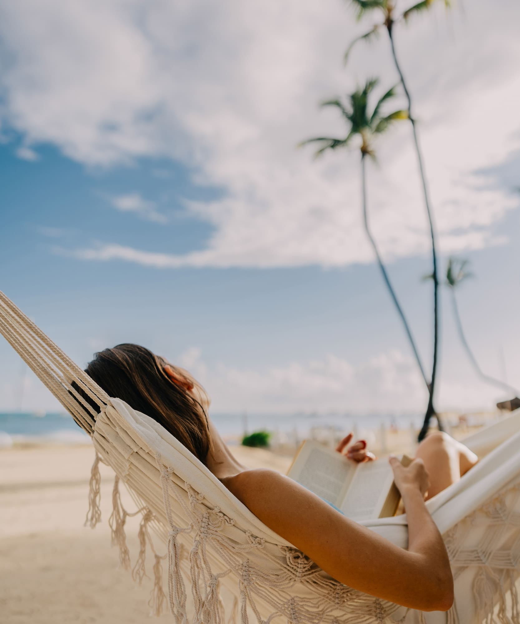 Woman reading book in hammock on tropical beach with palm trees.