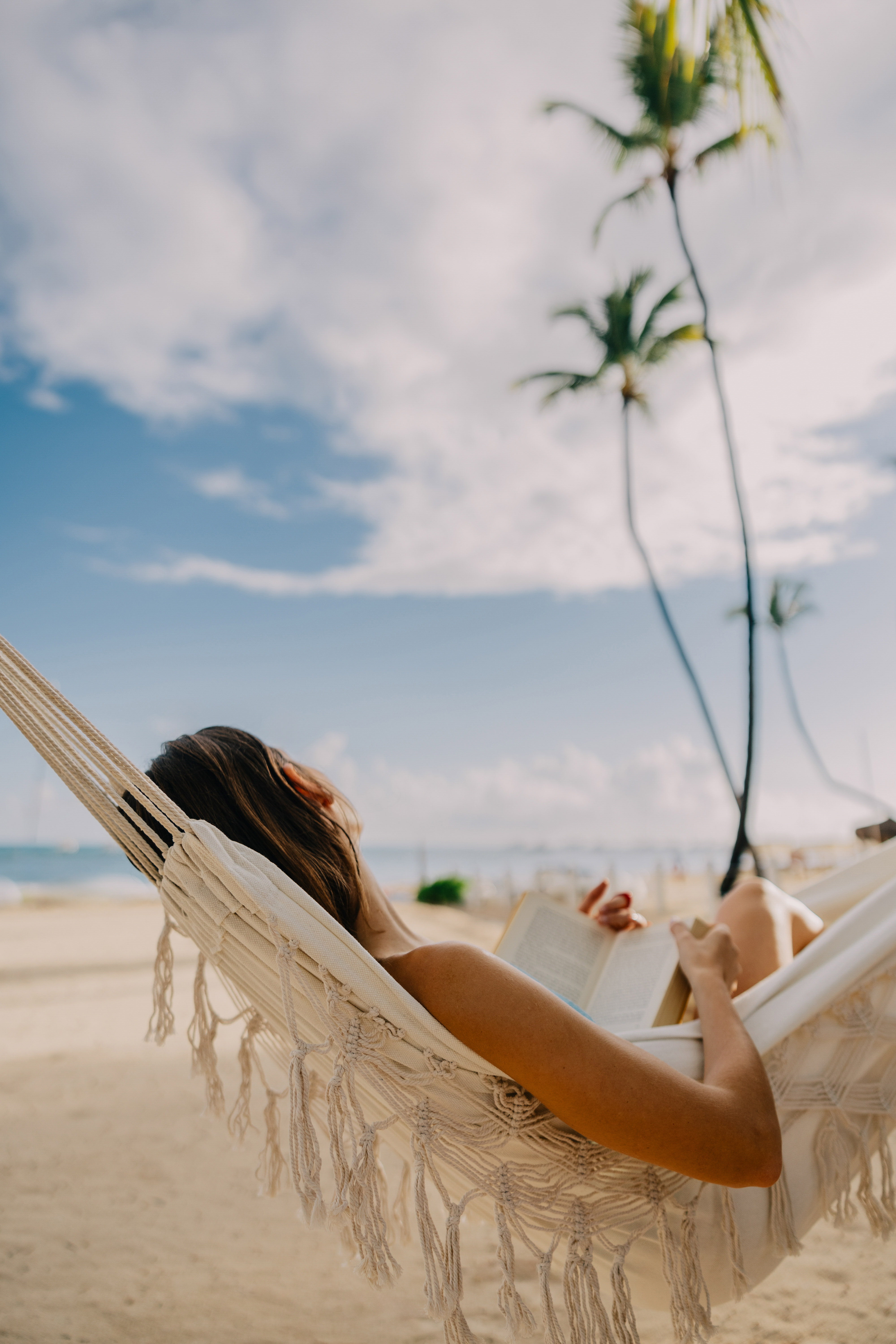 Woman reading a book in a hammock on a sunny beach with palm trees.
