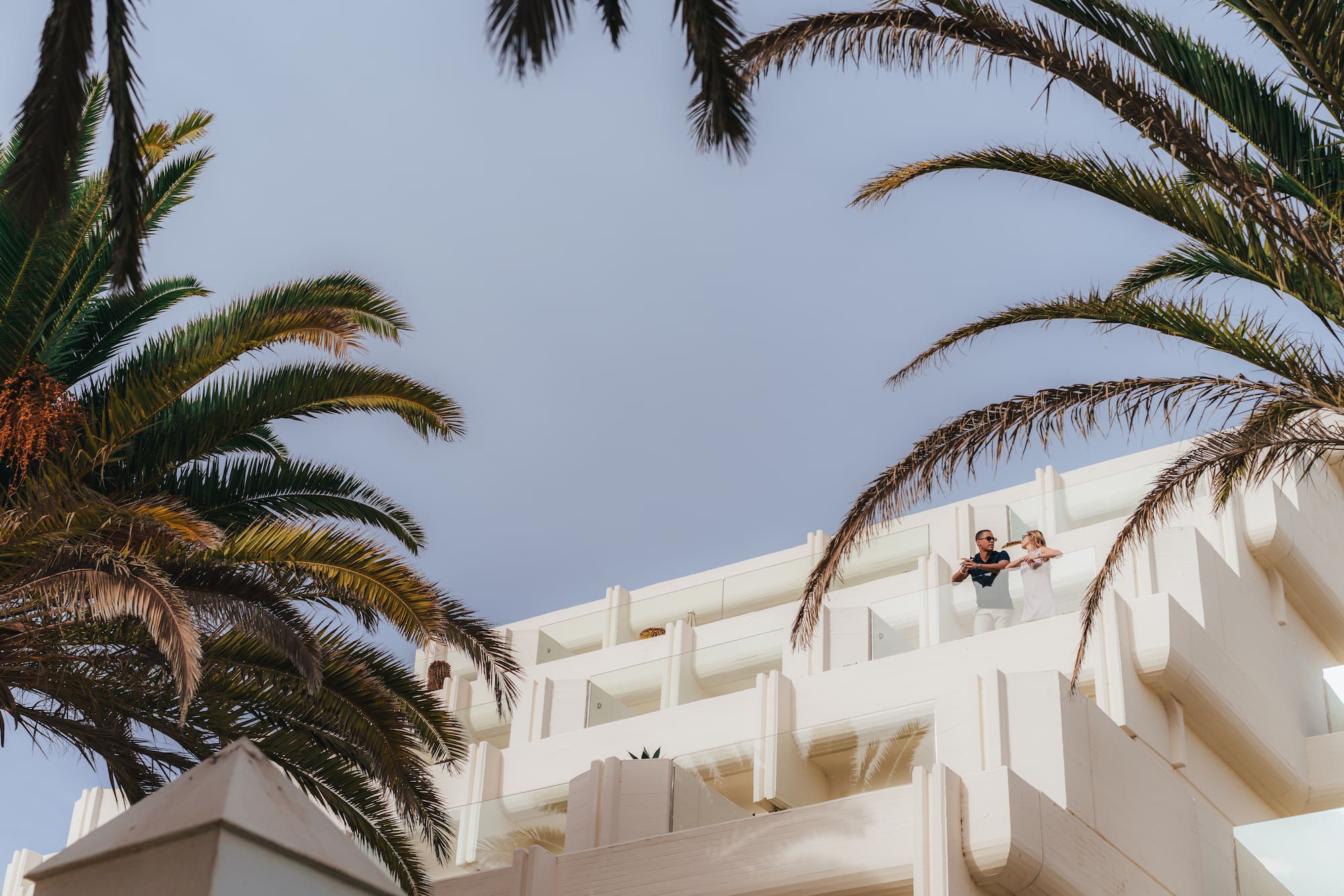 Pareja en balcón de hotel moderno blanco con palmeras, cielo azul.