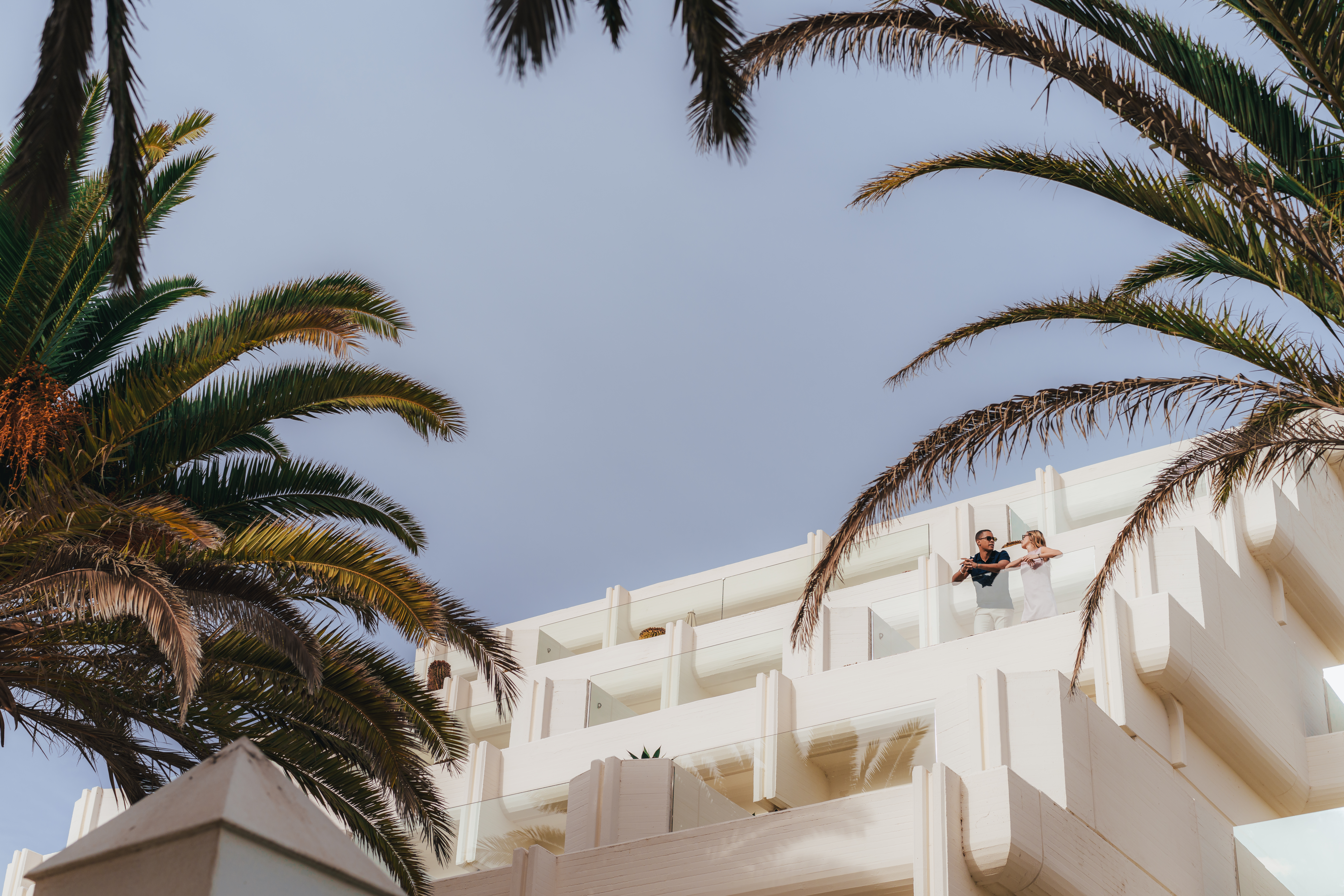 Pareja en balcón de hotel moderno blanco con palmeras, cielo azul.