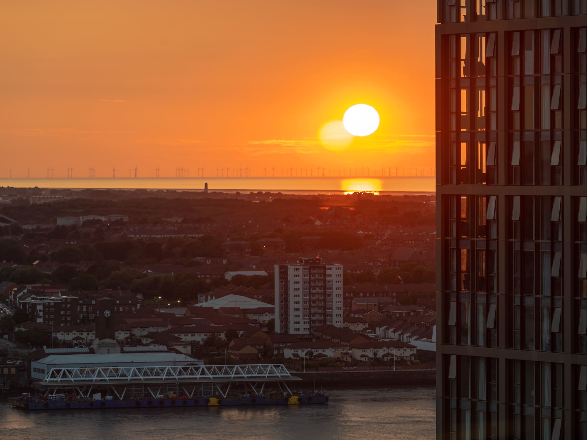 Orange sunset behind offshore wind turbines, urban riverfront, and skyscraper.