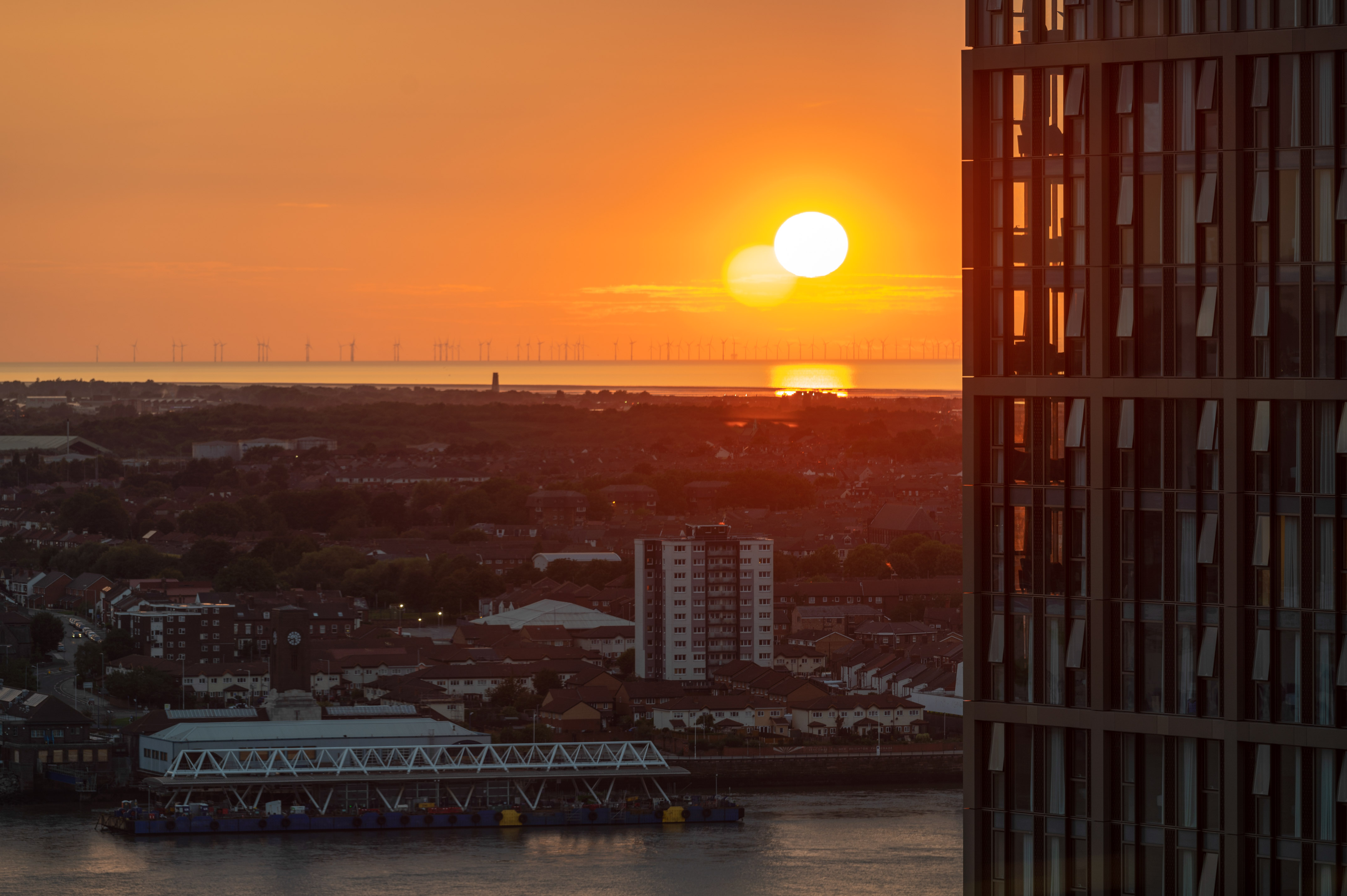 Orange sunset behind offshore wind turbines, urban riverfront, and skyscraper.