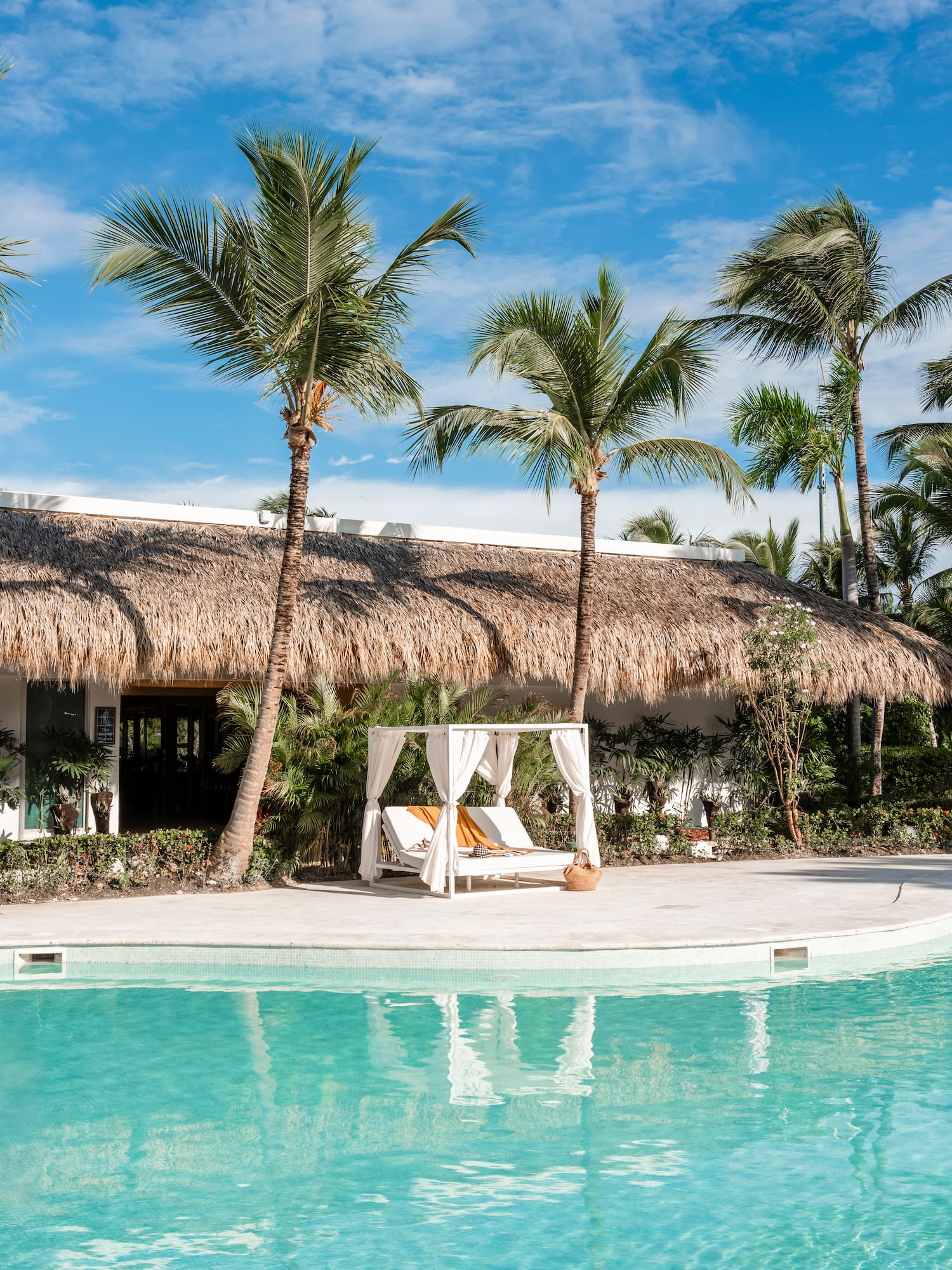 Tropical pool, thatch-roof building, palm trees, and poolside cabana under blue sky.