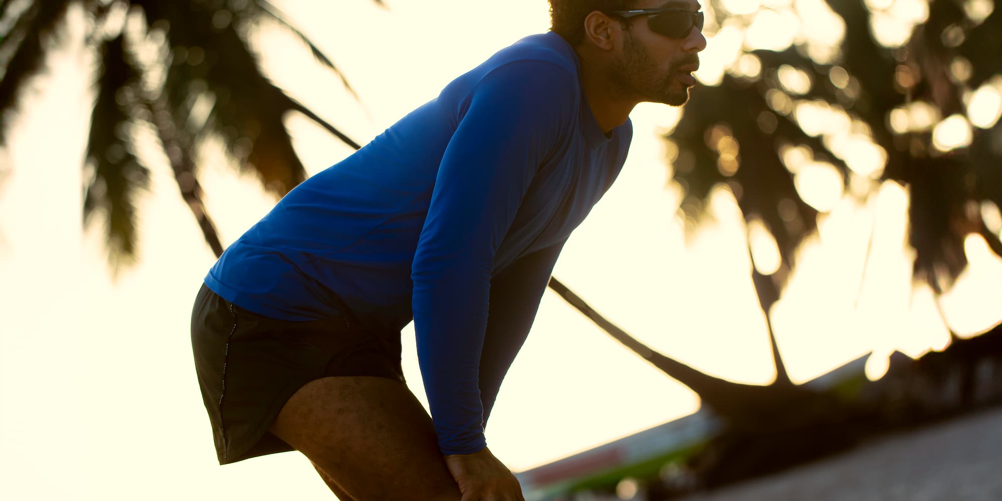 Male runner resting on sandy beach at sunset, silhouetted palm trees.