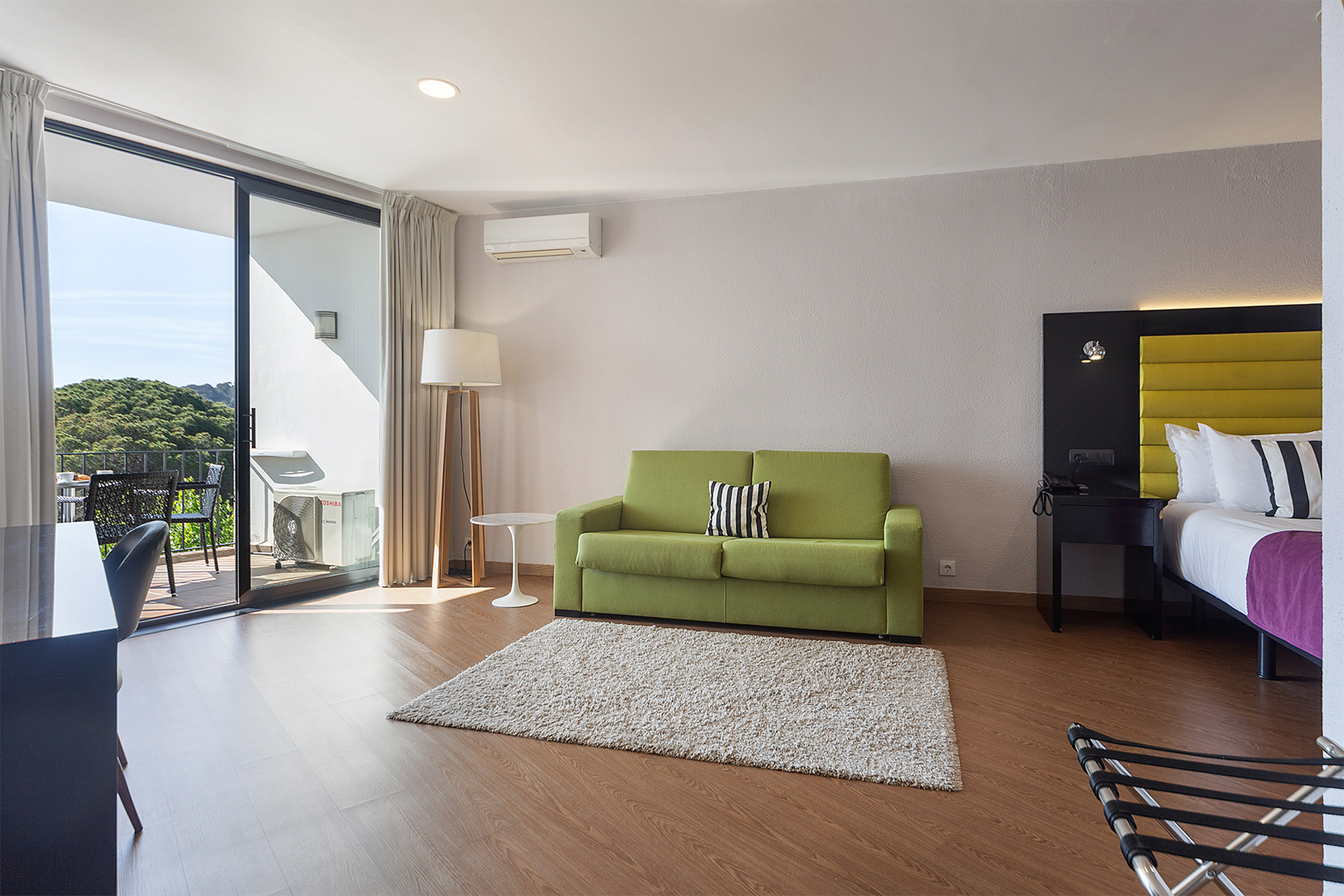 Modern hotel suite with green sofa, bed, and balcony overlooking green trees.