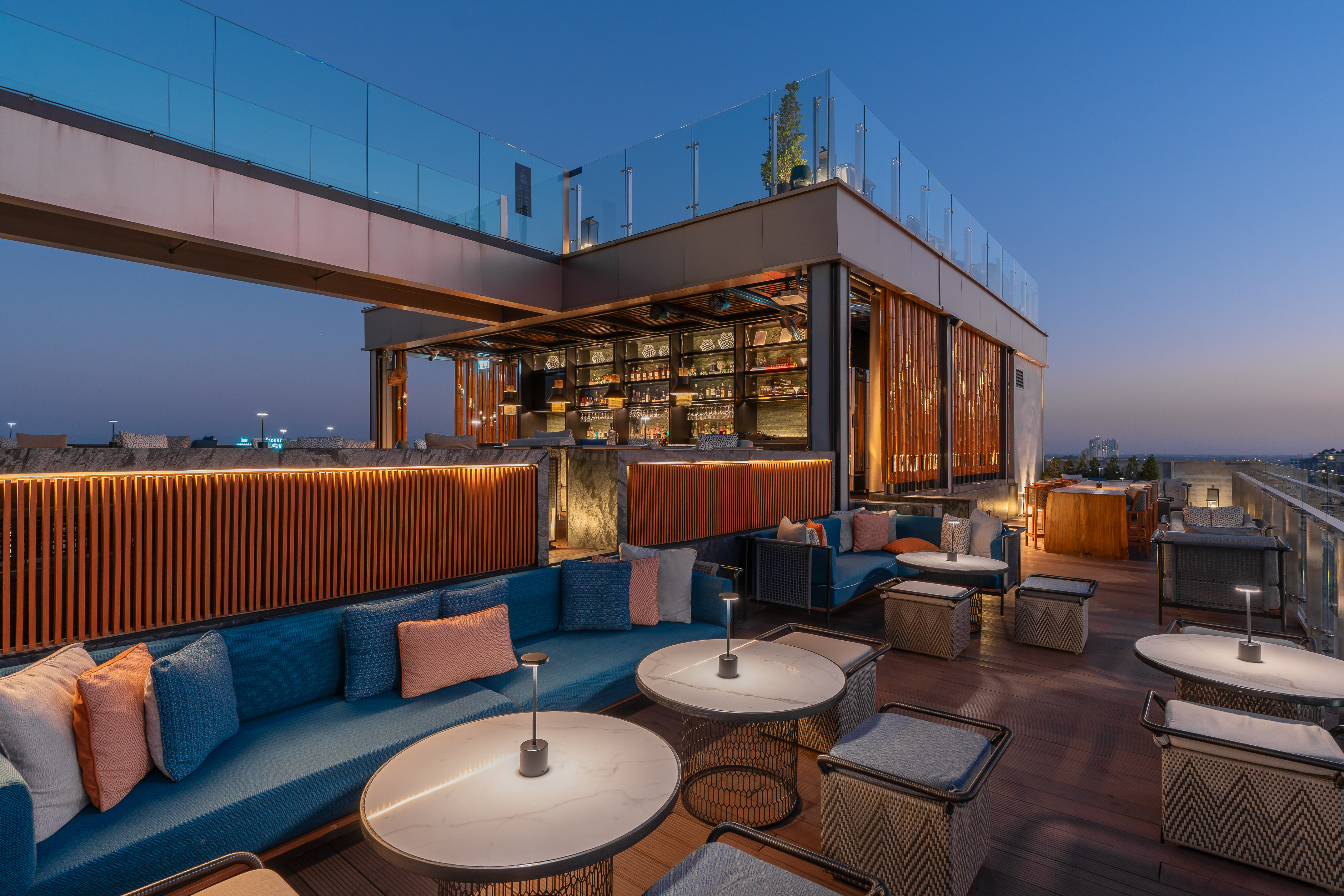 Modern rooftop bar at dusk with blue lounge seating, marble tables, and city views.