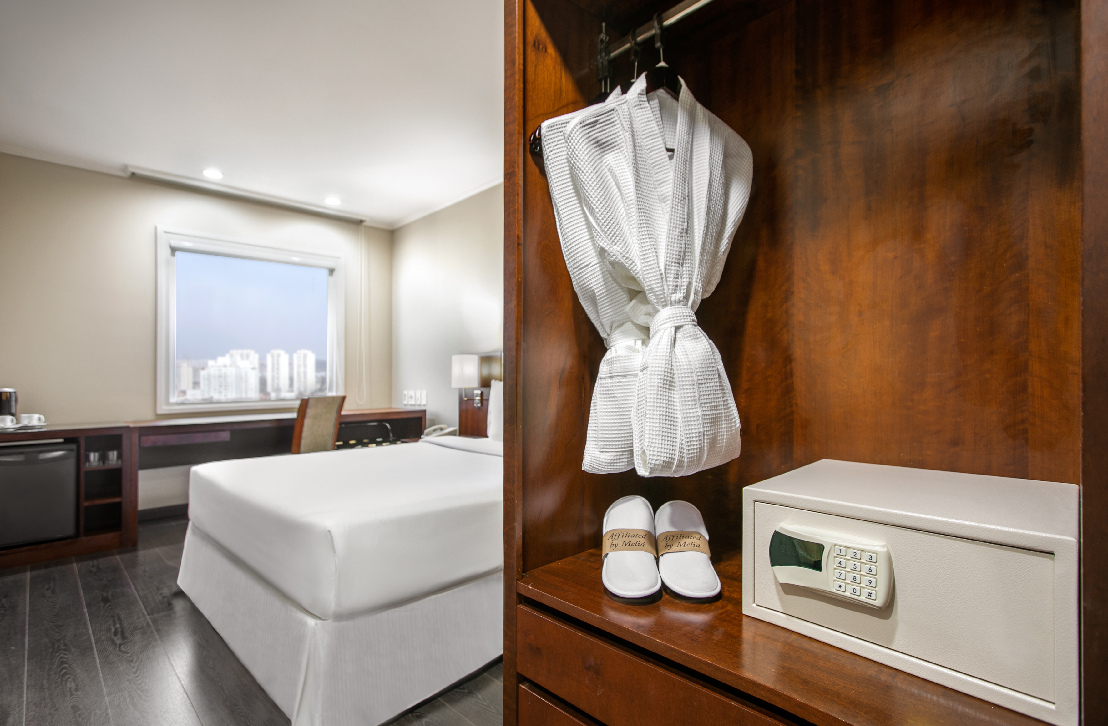 Hotel room with bed, city view, closet showing bathrobe, slippers, and safe.