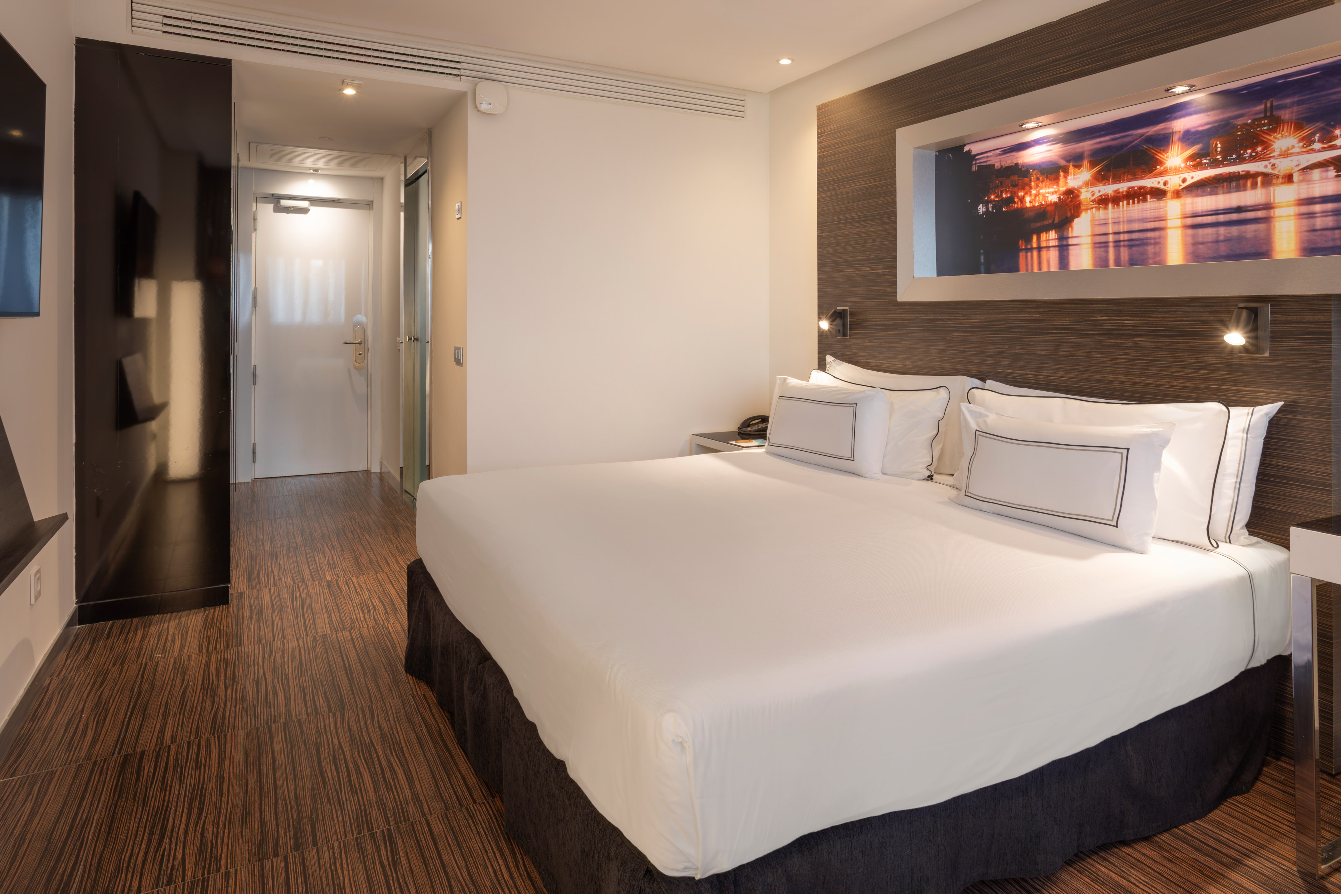 Hotel room with white king bed, dark wood floor, and illuminated cityscape art.