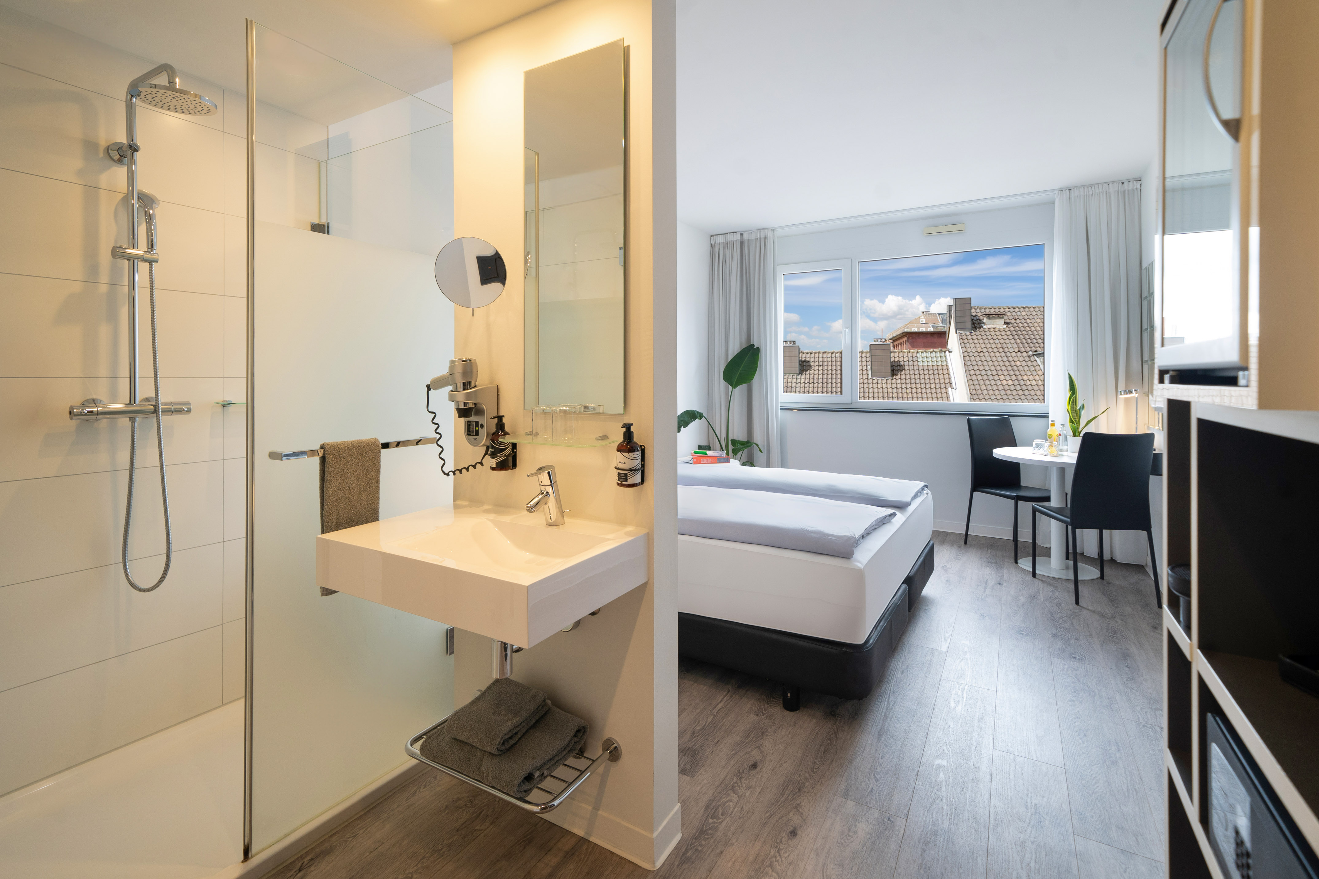 Modern hotel studio with open bathroom, bed, and city view window.