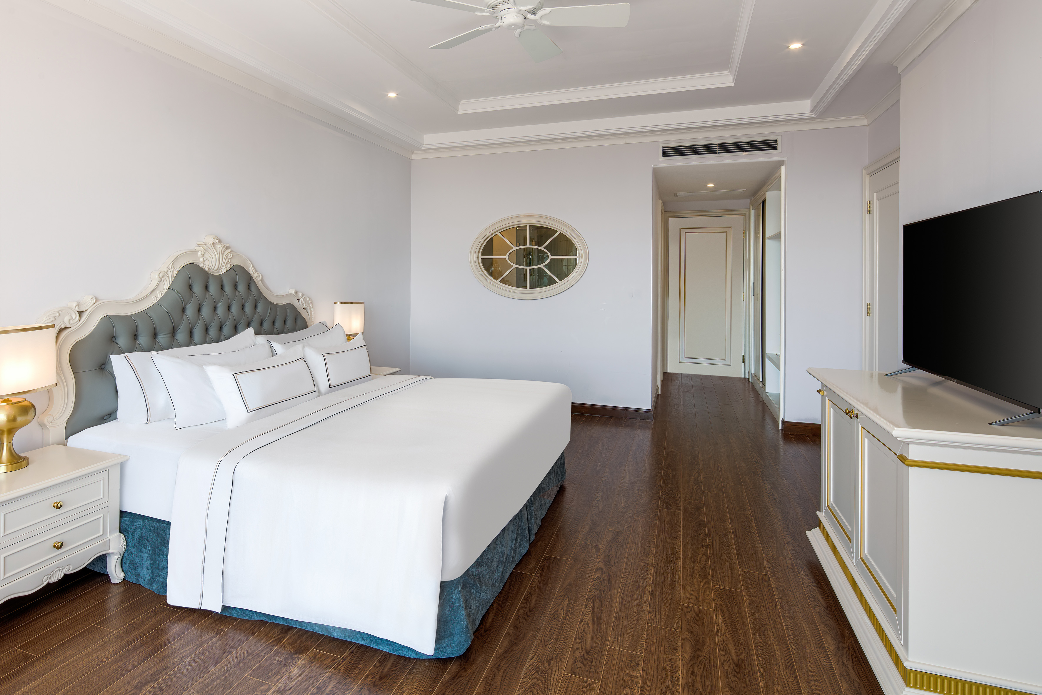 Elegant hotel bedroom with king bed, ornate headboard, dark wood floor, white walls.