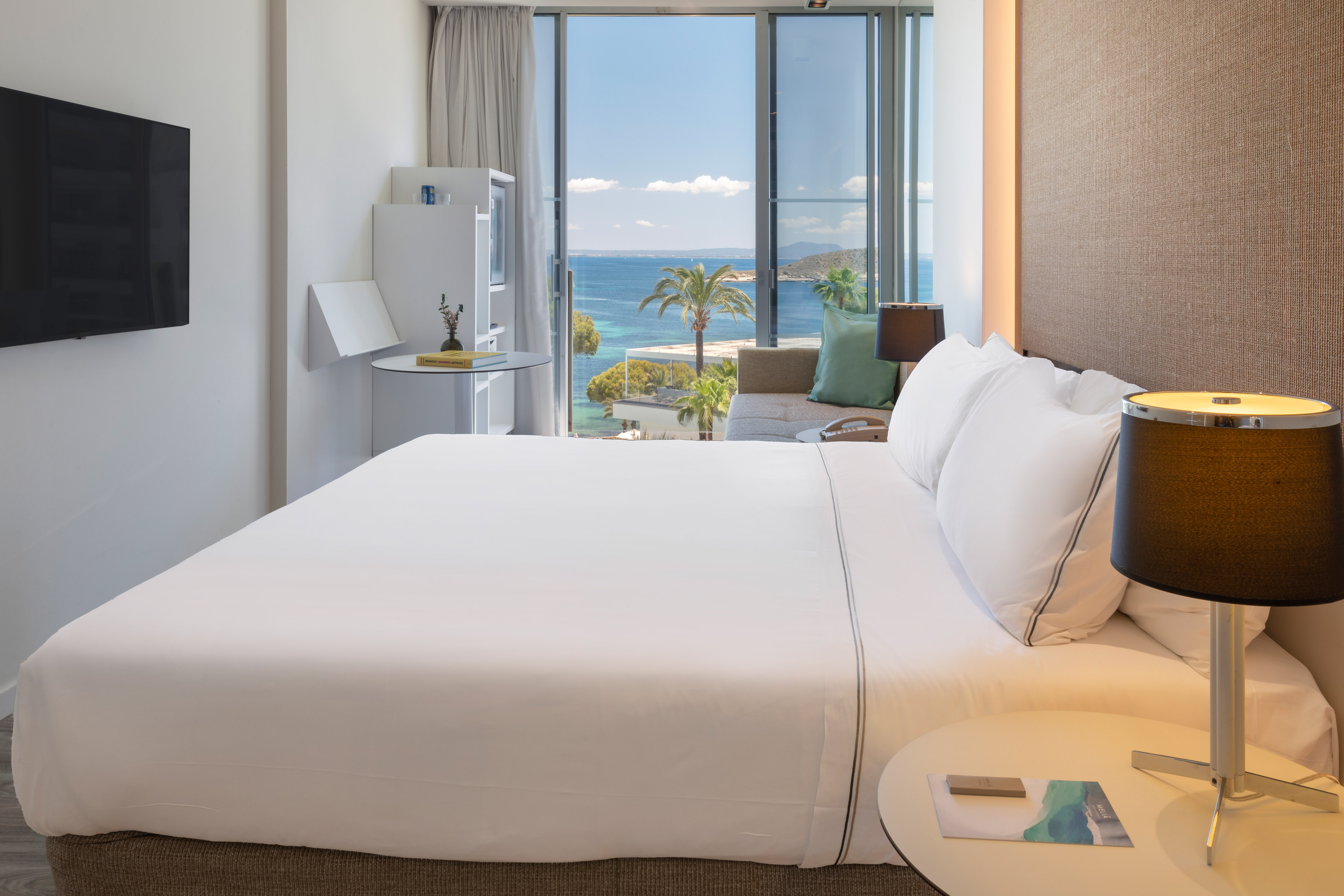 Modern hotel room with white bed, TV, and ocean view through large window.