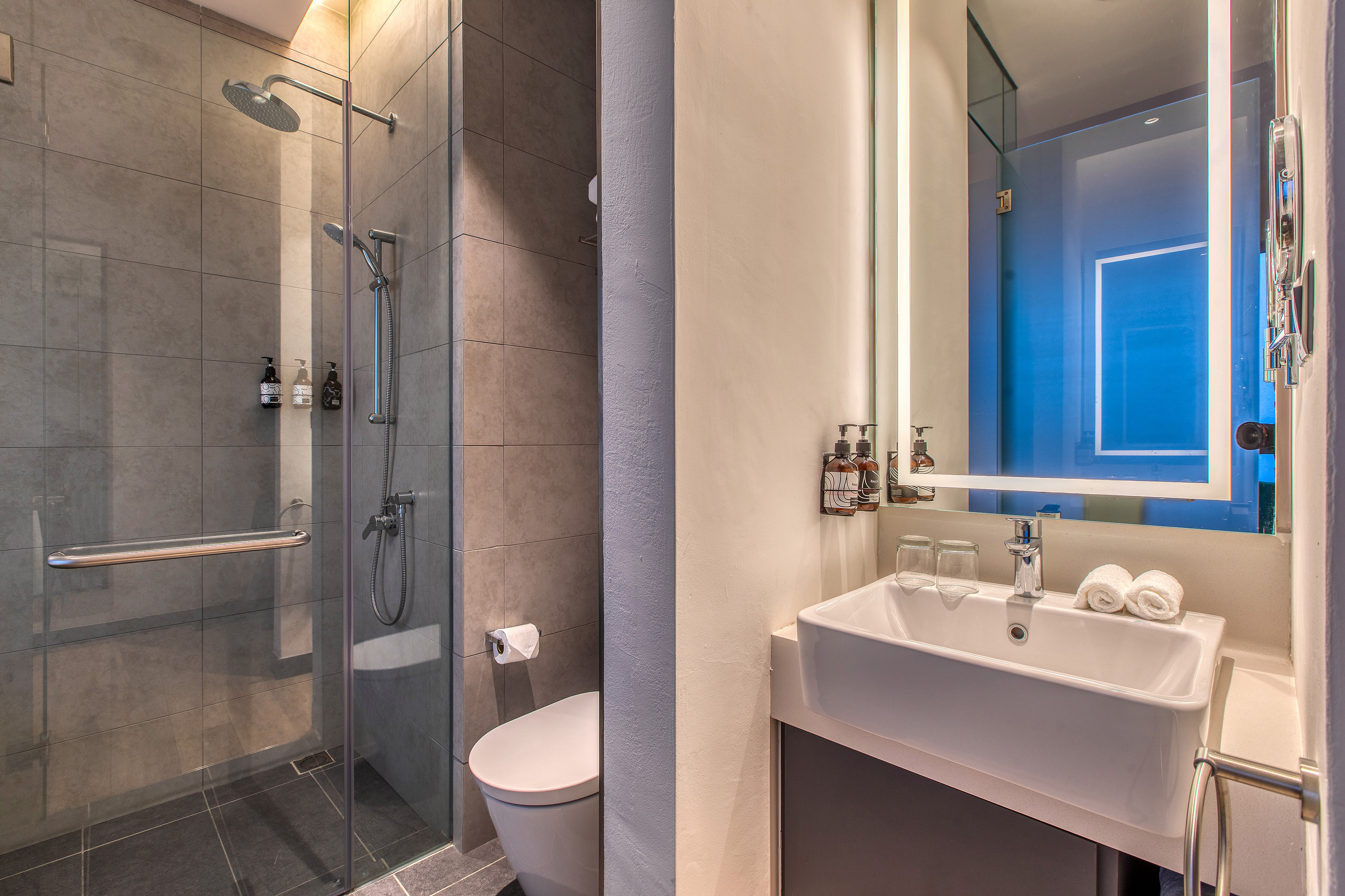 Hotel bathroom: glass shower, toilet, vanity, illuminated mirror.