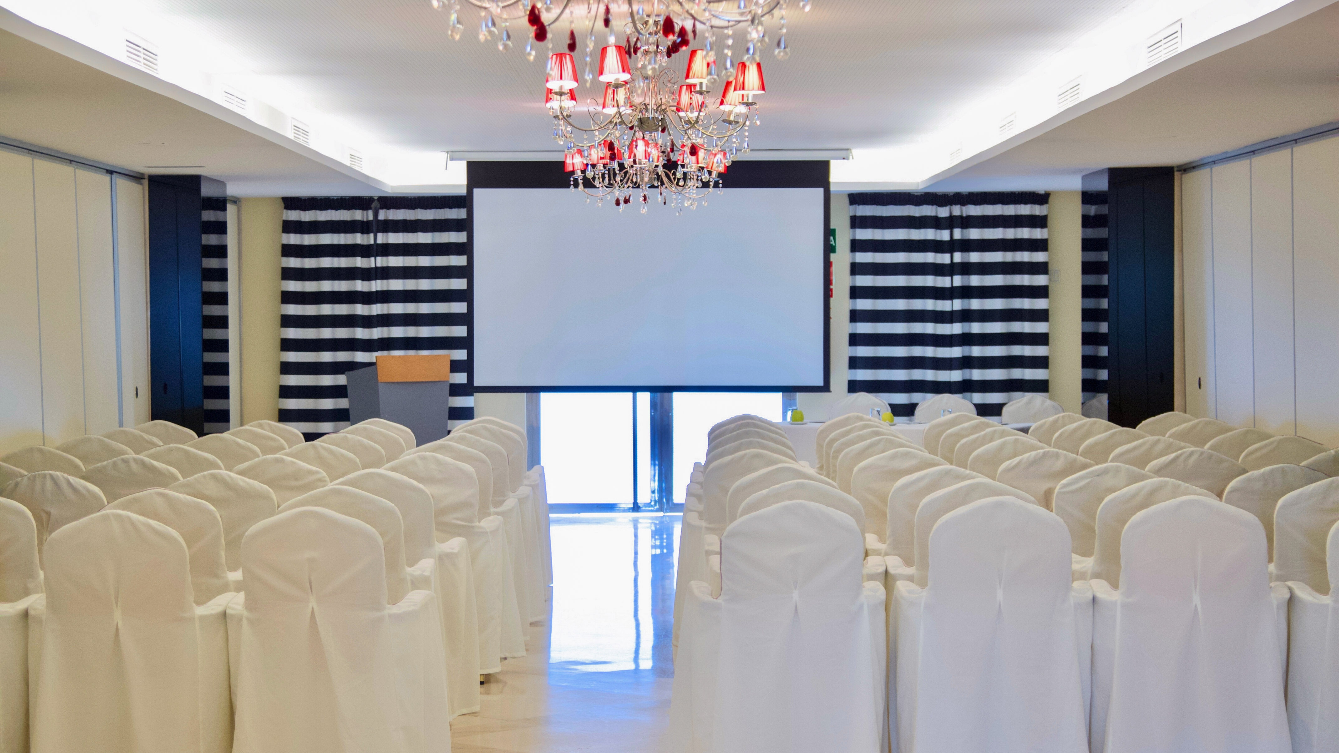 Conference room with white chairs, projector screen, podium, and chandelier.