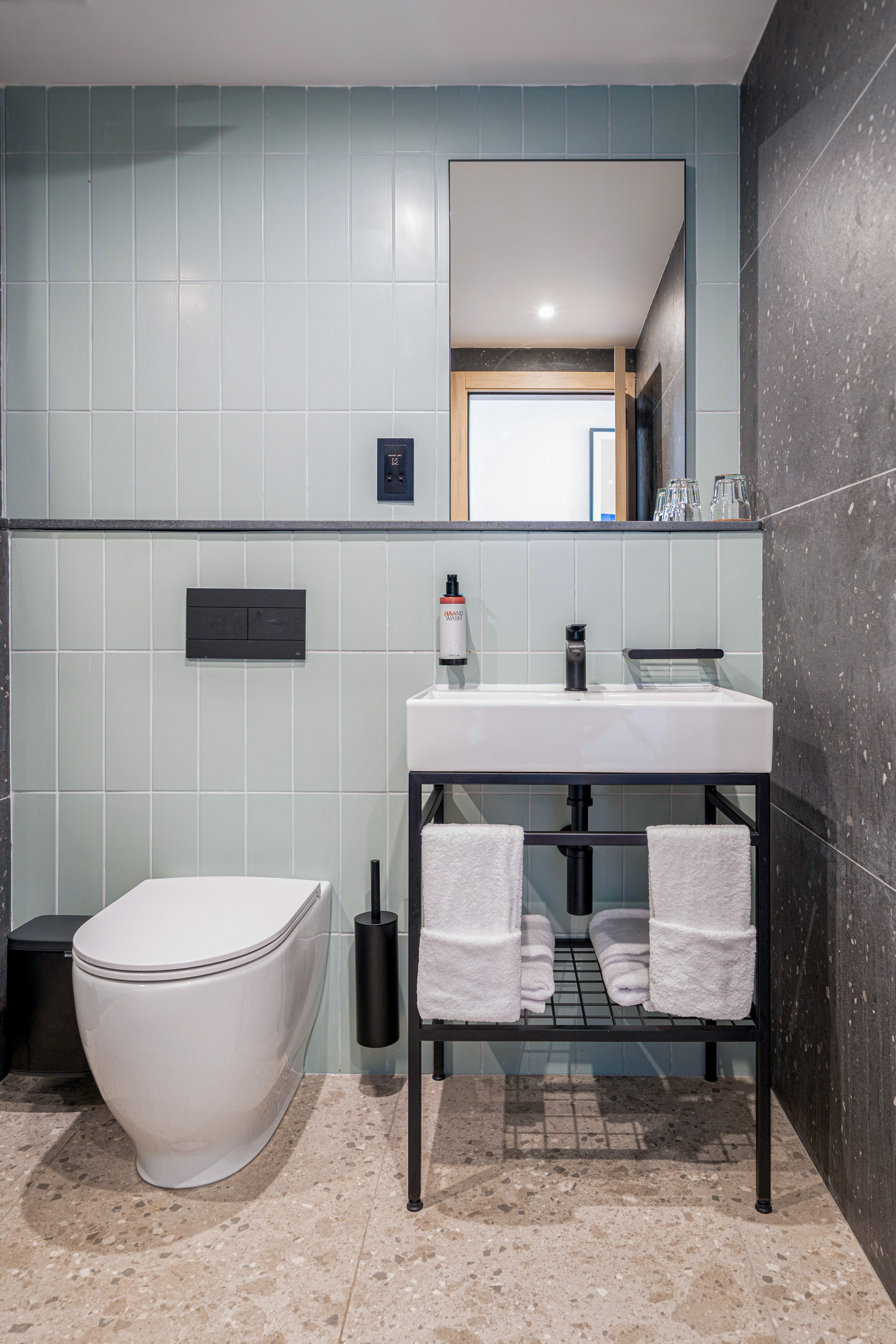 Modern bathroom: light blue wall tiles, white toilet, vanity, mirror, black fixtures.