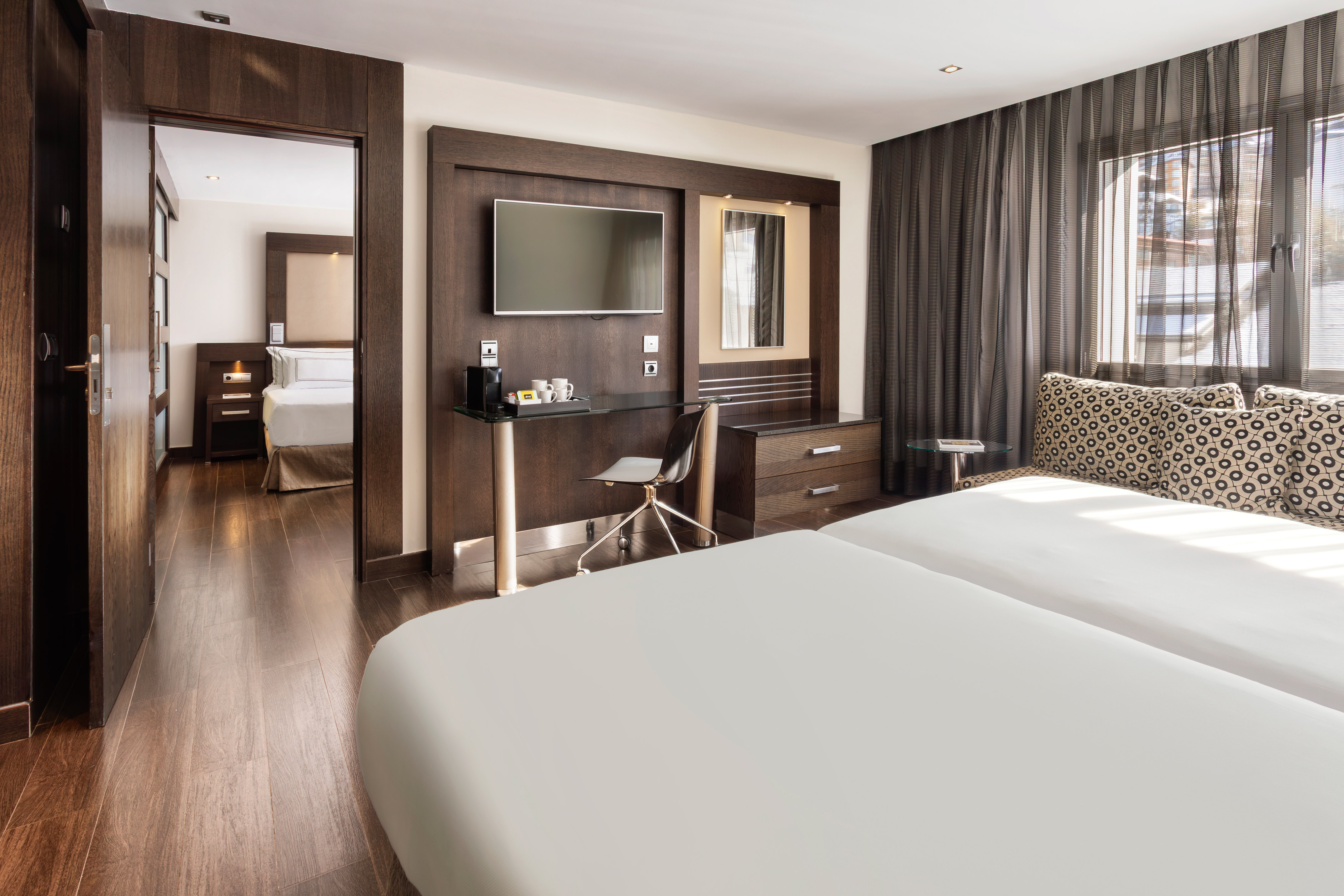 Modern hotel suite: twin beds, dark wood desk with TV, connecting bedroom.