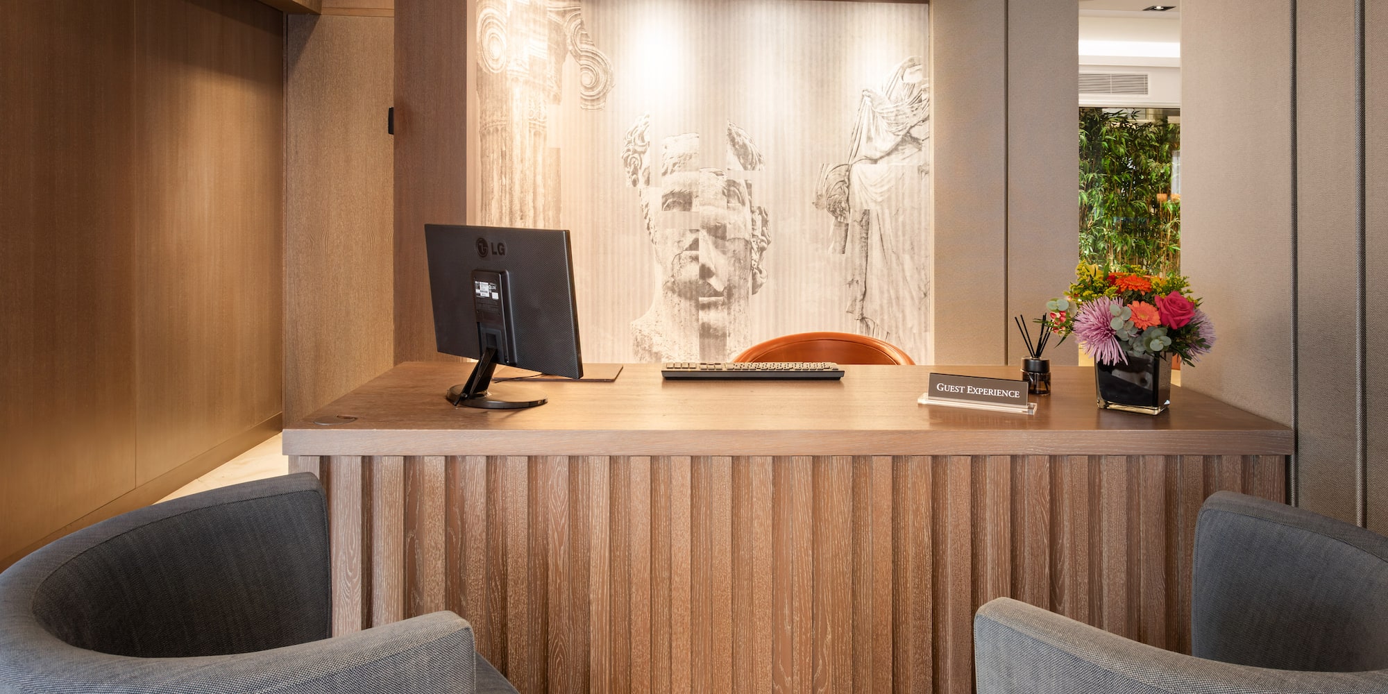 Modern hotel reception desk with wood details, classical mural, and "Guest Experience" sign.
