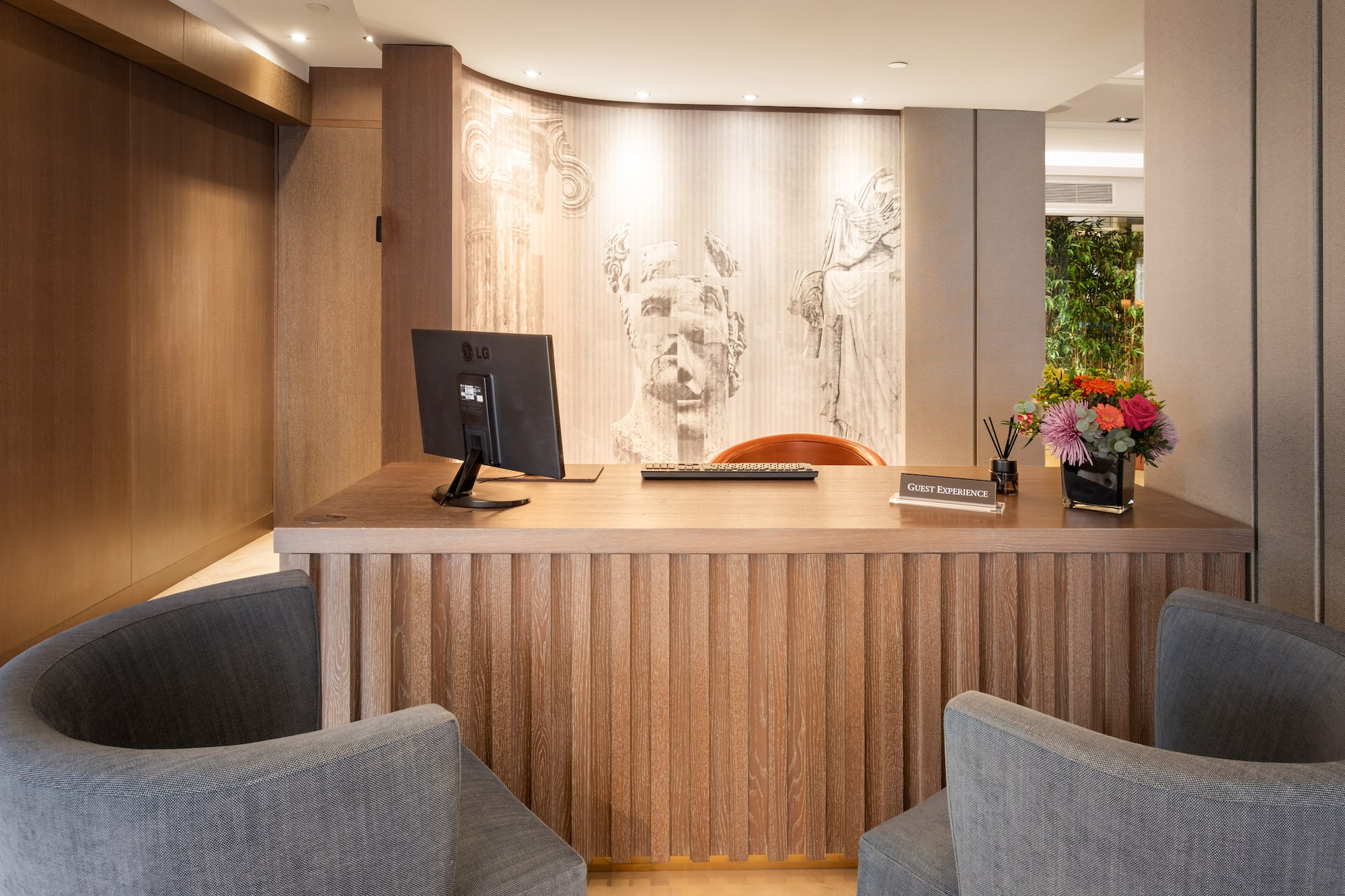 Modern hotel reception desk with wood details, classical mural, and "Guest Experience" sign.