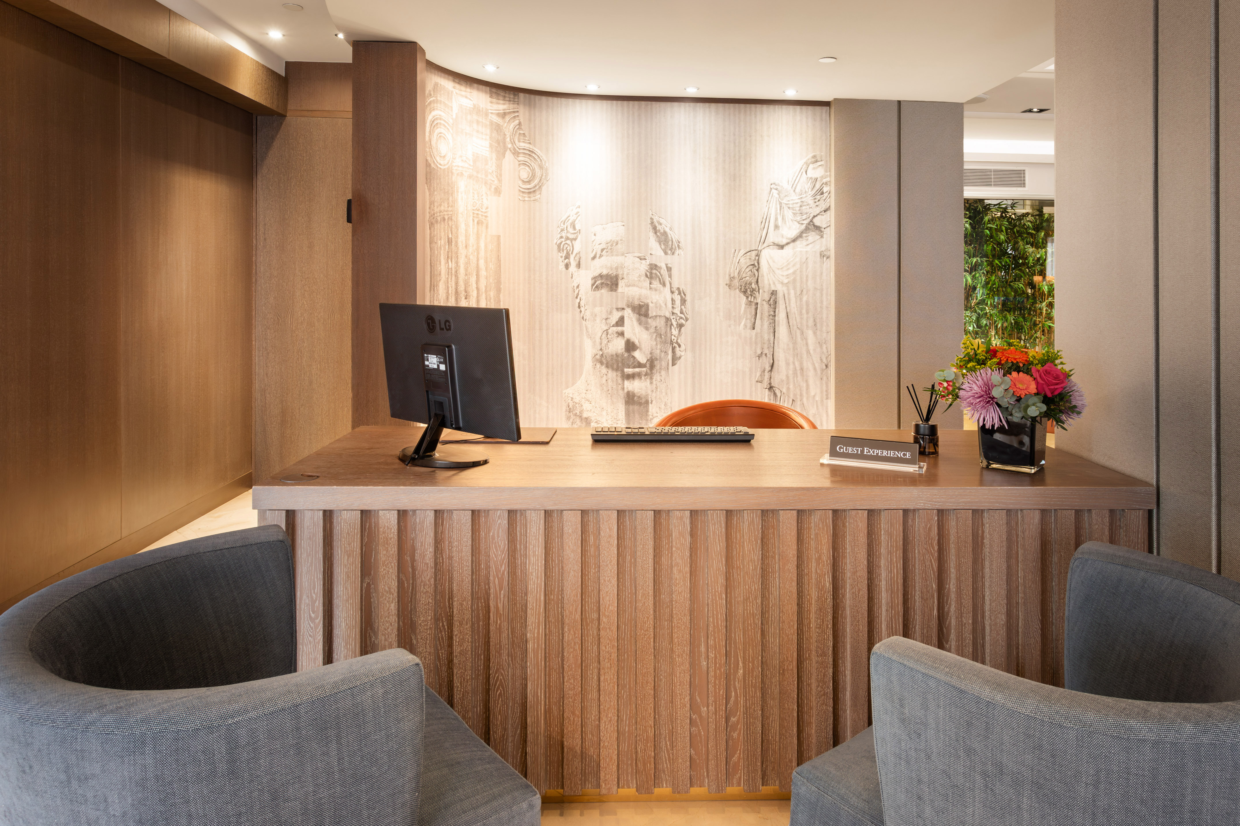 Modern hotel reception desk with wood details, classical mural, and "Guest Experience" sign.